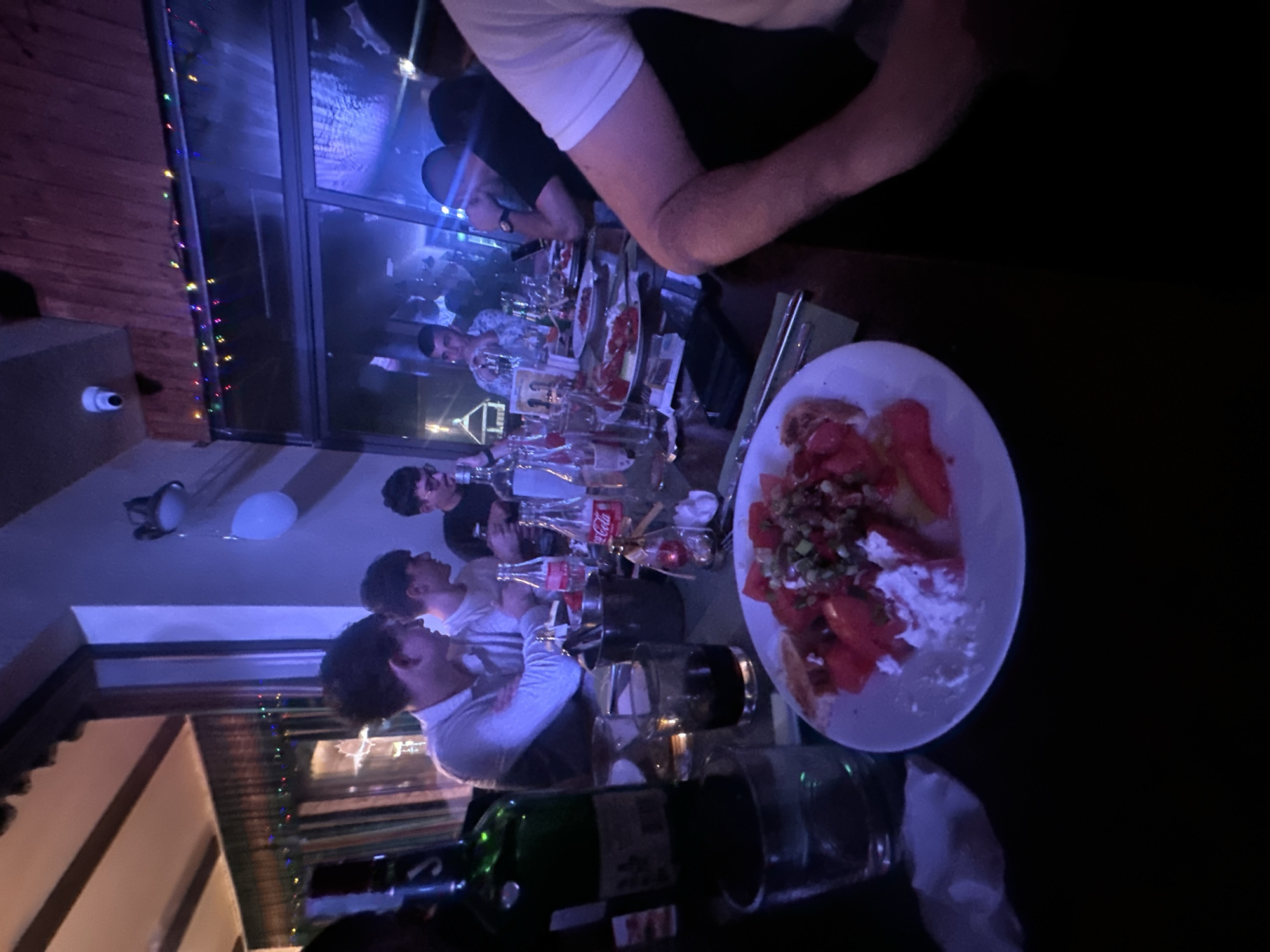 A dimly lit, indoor dining scene in Panagyurski kolonii, Bulgaria, shows a group of people seated at a long table. The table is covered with bottles of wine, water, and Coca-Cola, glasses, and dishes of food, including a plate of what appears to be tomatoes, feta cheese, and herbs. People of varying ages are engaged in conversation. A balloon and security camera are visible on the ceiling. Christmas lights adorn the window.