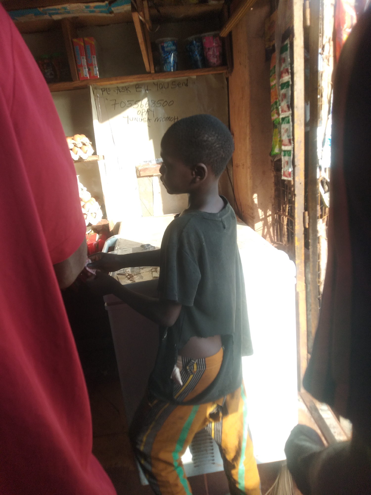 A young, dark-skinned child stands in what appears to be a small shop in Ejule, Nigeria. The child wears a dark green t-shirt and yellow pants with green stripes, and there are rips in the shirt and pants. The child is interacting with someone or something out of frame, towards the left. Behind the child is a white board with writing on it that reads "Please ask bil you send 7055668500 opayi tunusa momoh." There are shelves on the left holding various items, and sunlight pours in from an open doorway on the right. A red object is visible in the left of the frame and a dark object is on the right of the frame, both partially obscuring the scene.