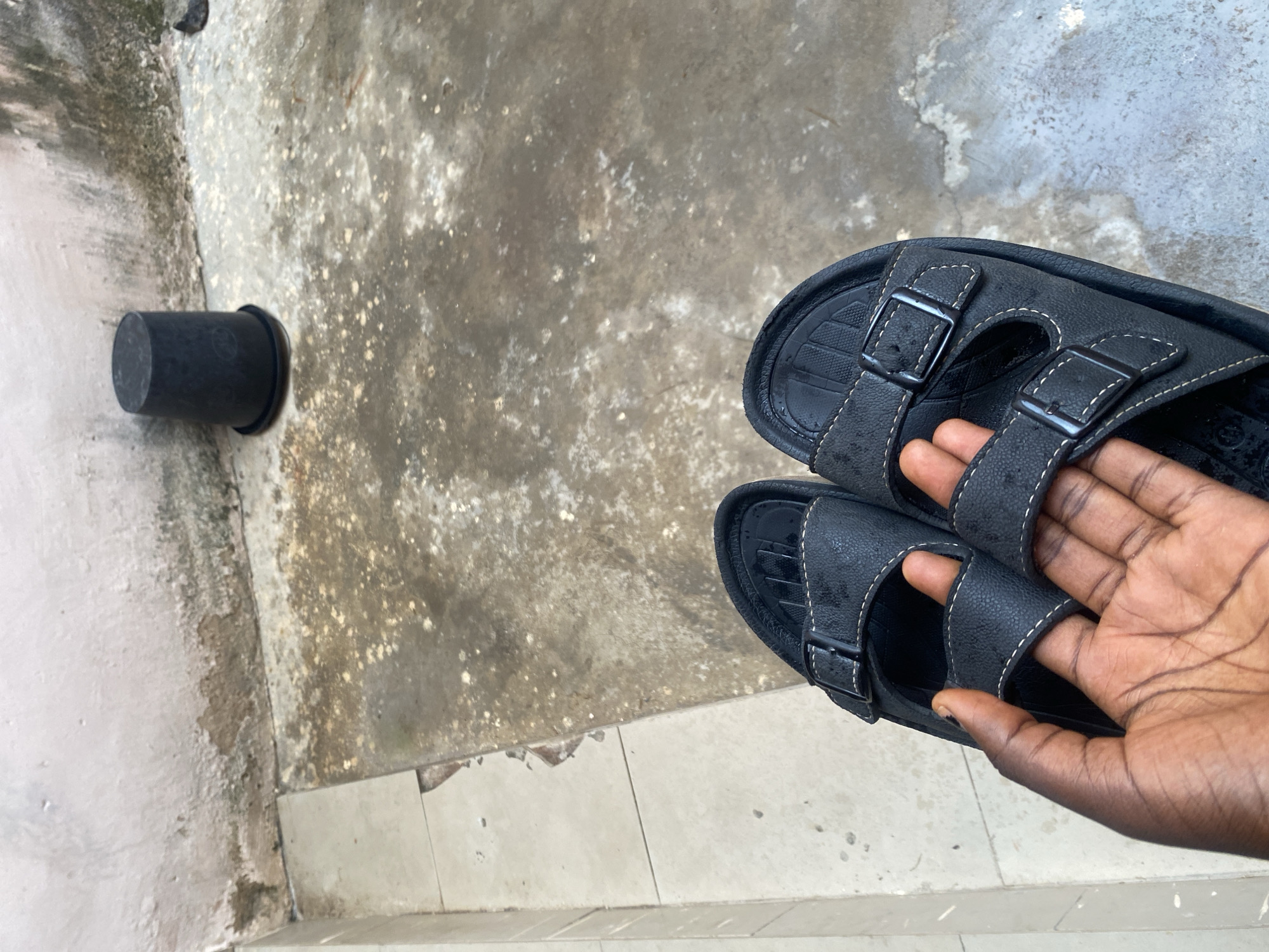A pair of black sandals is held by a person's hand, with the sandals positioned on a textured, greyish-white tiled surface. The sandals have two straps with buckles, and the stitching is white. To the left of the sandals, on a rougher, stained concrete surface, is a black cylindrical object, possibly a pot or a container. The scene appears to be outdoors, given the natural lighting and the weathered appearance of the surfaces. The overall impression is of a casual, everyday setting. There is no visible text in the image.