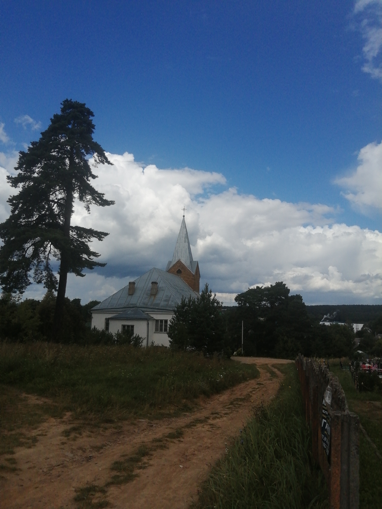 The image shows a rural landscape with a church and a tall pine tree in the foreground. A dirt road leads towards the church, which has a gray roof and a pointed steeple topped with a cross. The church appears to be a white building with some red brick visible near the steeple. The sky is blue with scattered white clouds. The surrounding area is covered in green grass and trees. In the distance, there is a line of trees and some buildings. The scene is likely in Slonim, Belarus, given the architecture and landscape. There are no people visible in the image. The overall impression is one of a peaceful, idyllic countryside scene.