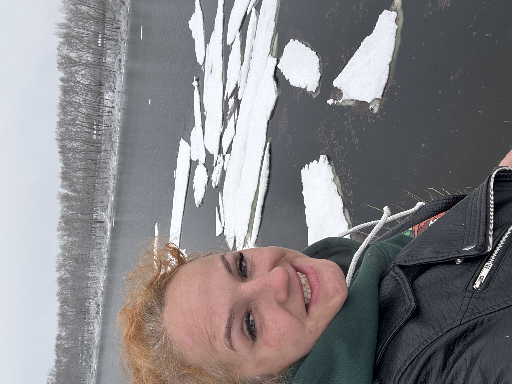 A selfie shows a smiling woman with light skin, blue eyes, and curly reddish-blonde hair. She is wearing a black leather jacket over a green hoodie. In the background, there is a body of water covered in ice floes. A tree-lined bank is visible on the far side of the water. The scene appears to be Chaikovsky, Russia.