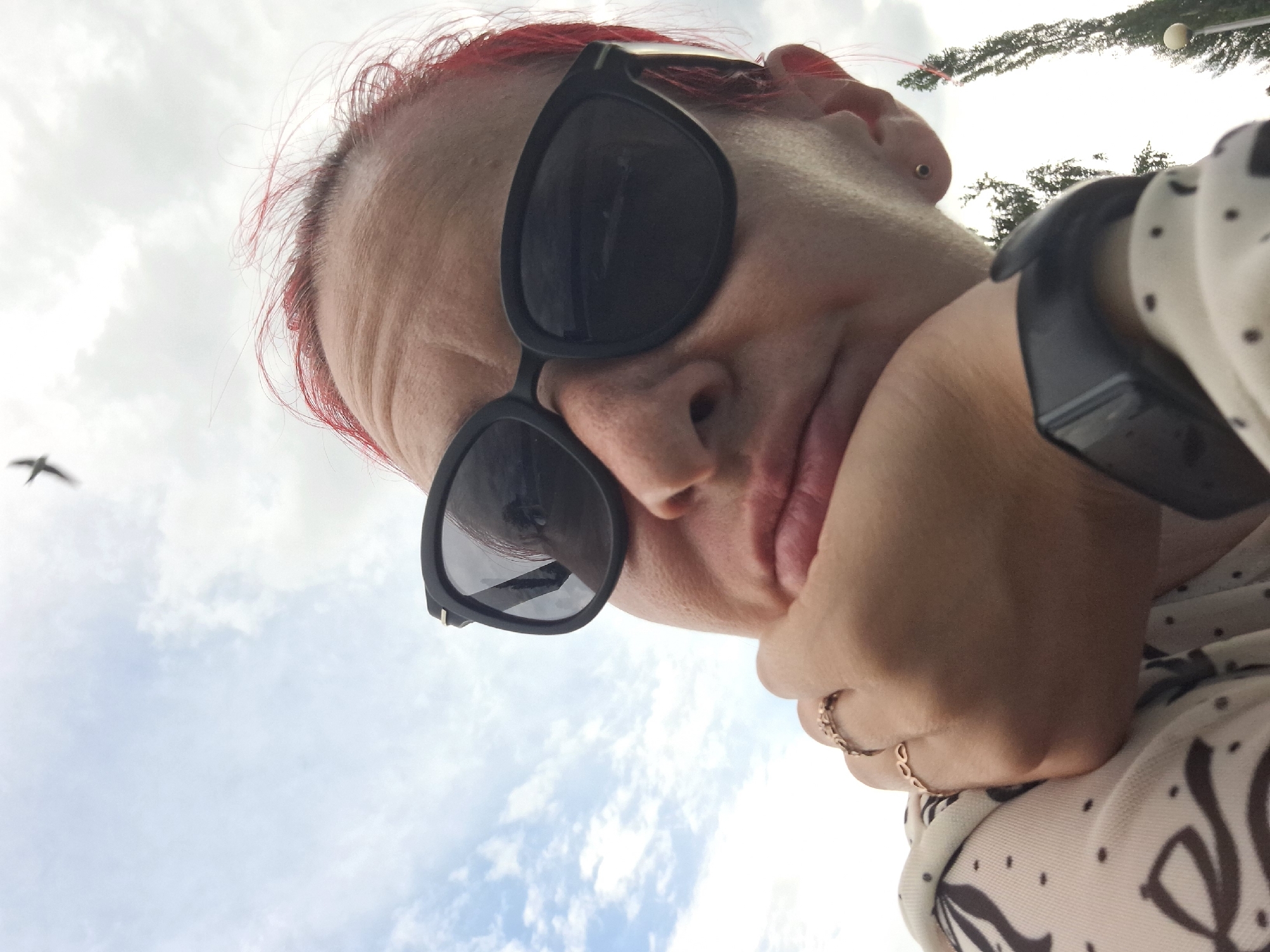 A low-angle, close-up selfie shows a person with red-dyed hair wearing black sunglasses and a light-colored, patterned shirt. The person's chin rests on their hand, which is adorned with two thin, gold-colored rings. A smartwatch is visible on the person's wrist. In the background, the sky is cloudy with a small bird flying overhead. Two tall trees are visible on the right side of the frame. The location is Atamanovka, Russia.