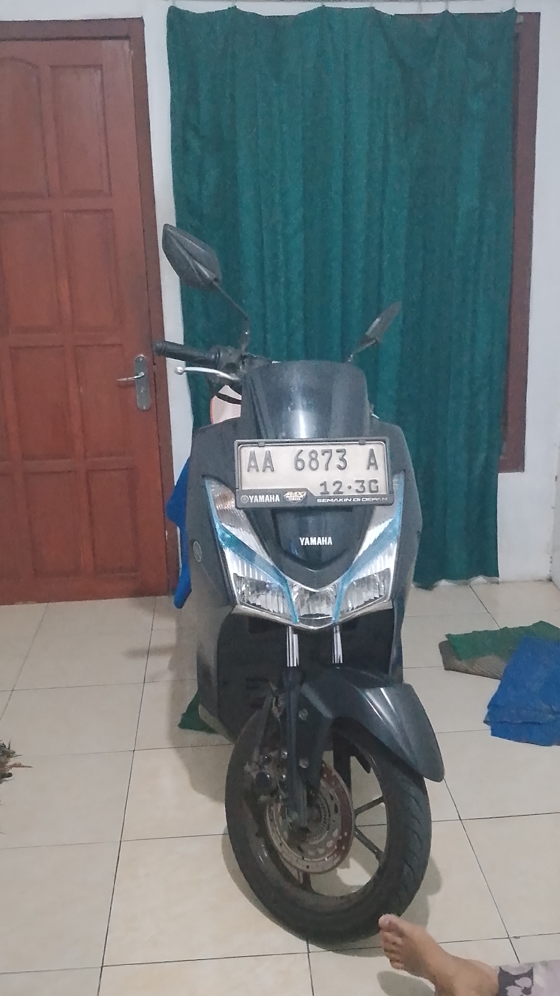 An eye-level medium shot shows a dark gray Yamaha scooter parked indoors. The scooter is facing the camera, with its front wheel slightly angled to the left. The license plate, which is white with black lettering, reads "AA 6873 A" and below it "12-30 SEMAKIN DI DEPAN". The scooter has blue accents on its headlights. To the left of the scooter is a dark brown wooden door with a silver handle. Behind the scooter is a dark green curtain. The floor is tiled with light brown and beige squares. In the bottom right corner of the image, a person's bare foot is visible. The overall lighting suggests an indoor setting with artificial light.