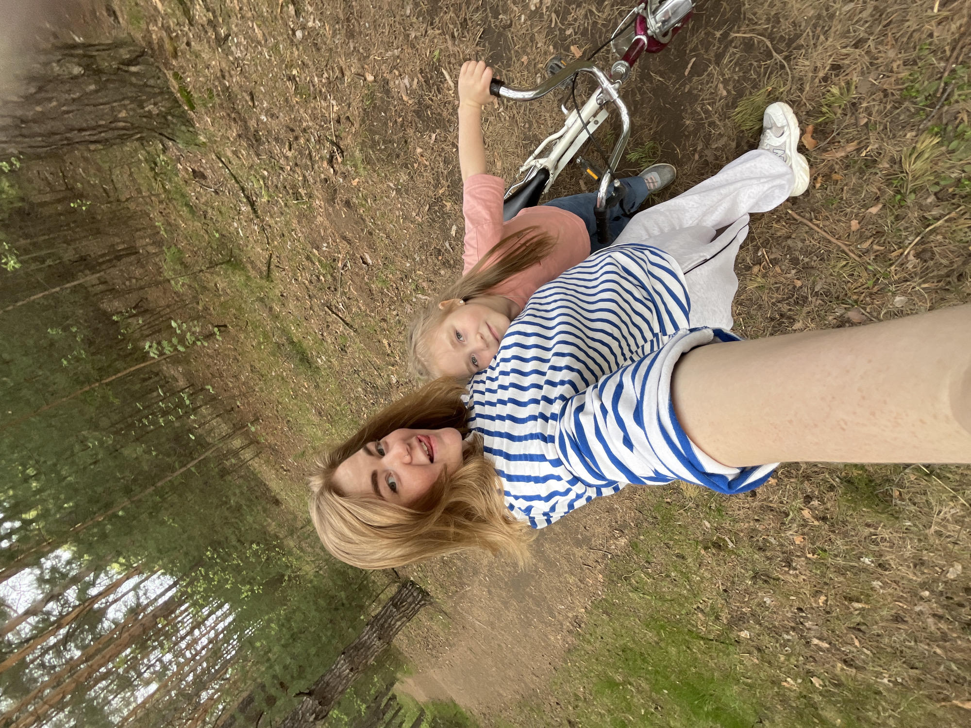The image captures a woman and a young girl in a forest setting in the city of Chaikovsky, Russia. The woman, wearing a blue and white striped shirt and light-colored pants, appears to be holding a camera or phone to take a selfie. Her blond hair is shoulder-length. The young girl, with blond hair and wearing a pink top, is positioned behind the woman, looking towards the camera. A bicycle is visible in the upper right corner of the image. The ground is covered with grass, leaves, and scattered branches. The background shows a forest with trees. The image is taken from a high angle, suggesting the photographer is standing.