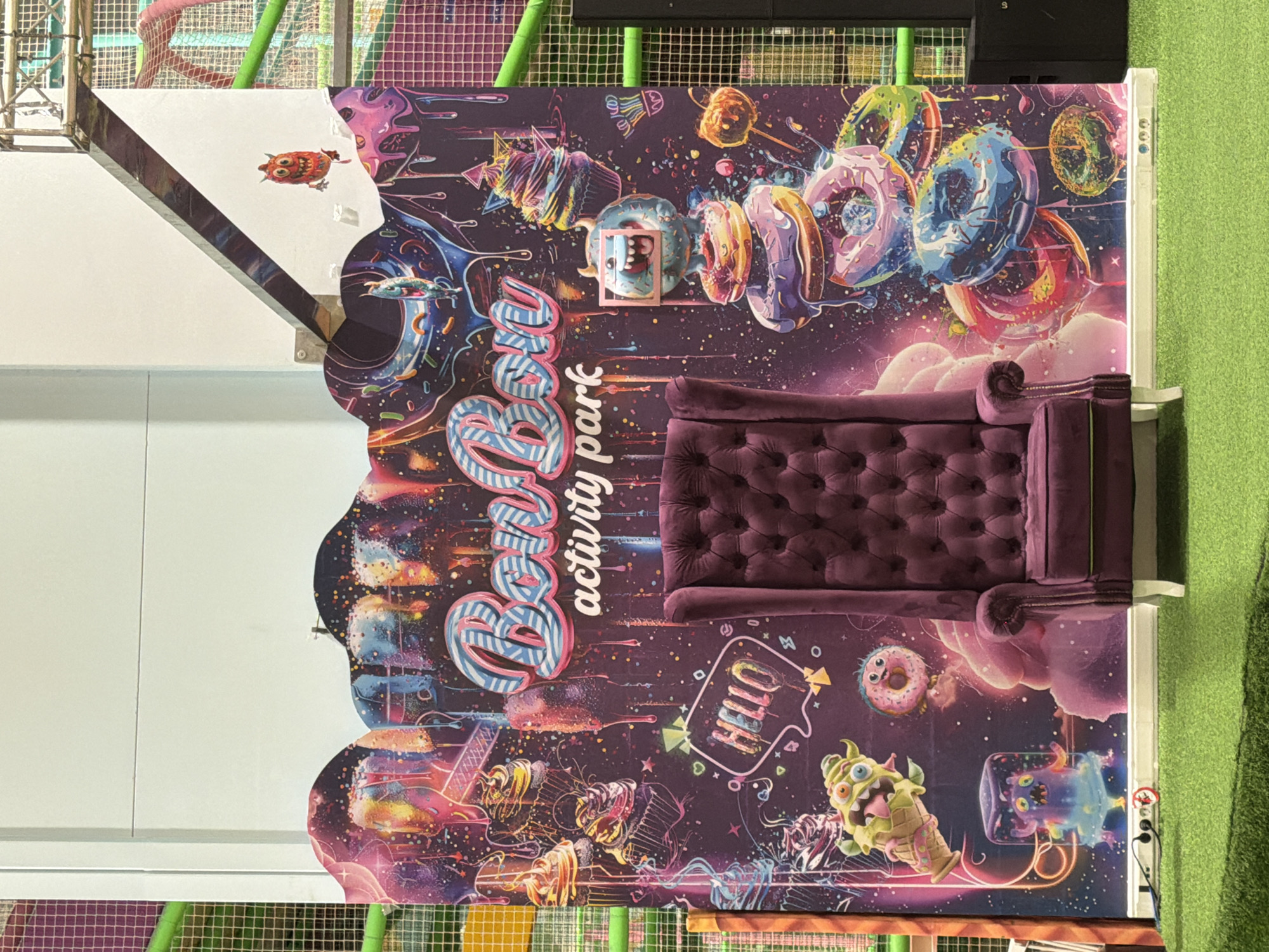 The image shows a large, colorful mural depicting a whimsical and fantastical scene with a space-like background filled with stars, nebulae, and glowing elements. Scattered throughout the mural are various cartoonish illustrations of donuts, cupcakes, and whimsical monsters. Prominently featured in the center of the mural is a plush purple armchair with a tufted back. The mural has a wavy, scalloped edge along the top.
On the left side of the mural, a small, orange, owl-like creature with large eyes is illustrated. To the right of this creature, a blue and white striped design forms part of the mural's text. Below this, the words "Boum" and "activity park" are visible in stylized fonts. The word "Boum" is depicted in large, playful, rounded letters with blue outlines and white stripes. "activity park" is written below in a smaller, white, sans-serif font.
Further to the right, a speech bubble-shaped graphic contains the word "HELLO" in blocky, yellow letters with a green outline. More whimsical monsters are scattered across the mural, including one that appears to be made of ice cream in a cone, and another that is trapped in a clear, blue container.
The mural is positioned against a white wall, and a bright green carpeted floor is visible in the background. To the left of the mural, a section of a play structure with metal bars and colorful tubes can be seen, suggesting the mural is part of a children's activity park or play area. The overall mood of the image is cheerful and energetic, likely intended for children. There are no visible people in the image. The lighting appears to be bright and even, typical of an indoor setting. There are no obvious time of day or weather cues. The location is identified as Qarağandı city administration, Kazakhstan.