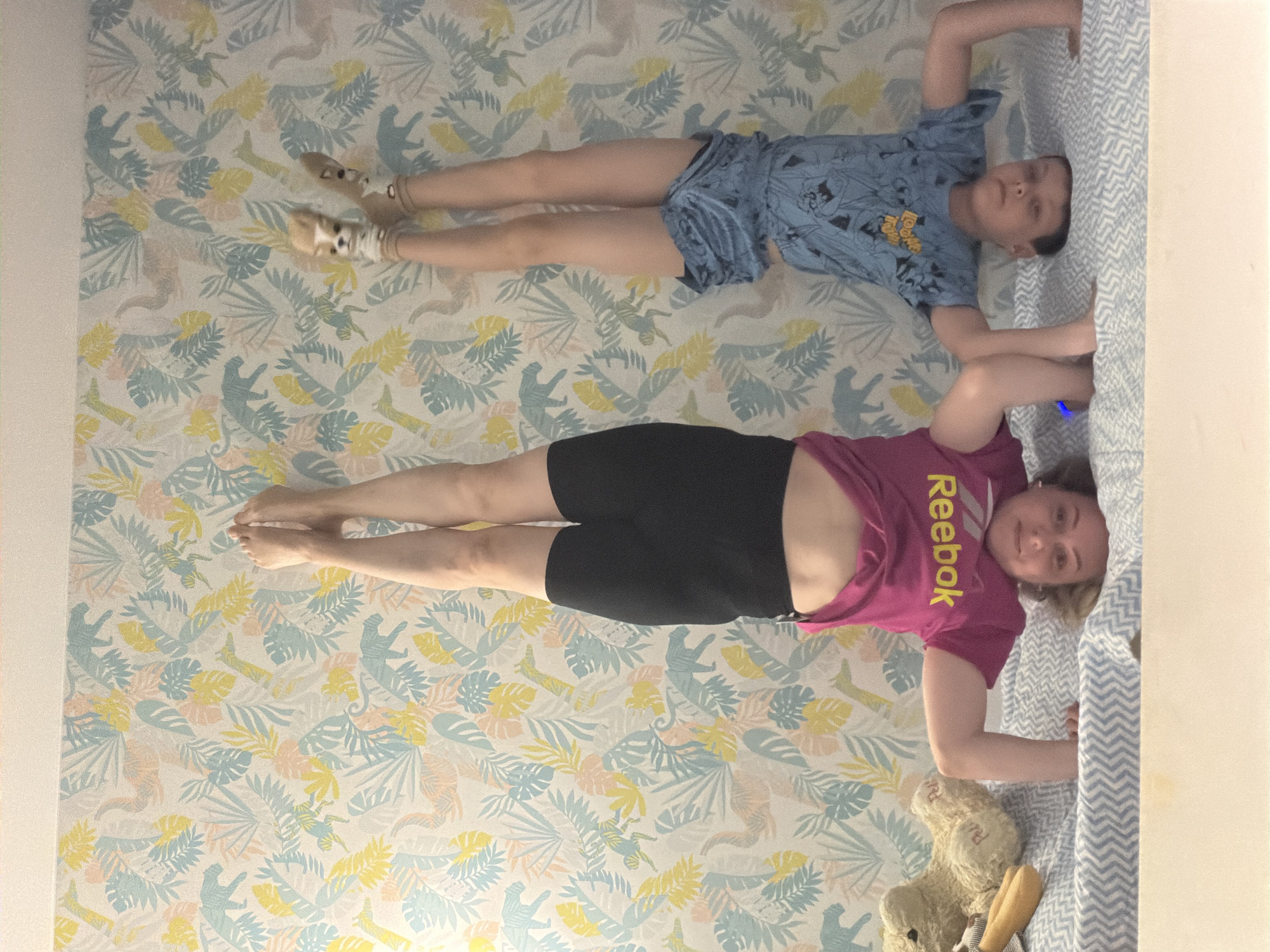 An overhead view captures an adult woman and a child lying side-by-side on a light-colored surface patterned with tropical foliage and animal silhouettes in Votkinsk, Russia. The woman, positioned in the lower half of the frame, wears a fuchsia t-shirt with "Reebok" printed in yellow and black shorts. She has fair skin and light hair, looking towards the camera with a slight smile. Her bare feet are visible towards the left.
Above her, a child, likely a boy, is dressed in a light blue t-shirt with a dark graphic print and blue patterned shorts. He has fair skin and dark hair, also looking towards the camera with a neutral expression. His feet are covered in light-colored socks with dark patterns. Both individuals have their heads oriented towards the right side of the frame, resting against a bed structure covered with a blue and white zig-zag patterned sheet.
Near the woman's feet, on the left, a light-colored teddy bear with a striped outfit is visible. The patterned surface features a repeating design of pale yellow, light blue, and peach-colored leaves, interspersed with silhouettes resembling monkeys and dinosaur-like creatures. The scene takes place indoors, conveying a relaxed atmosphere.