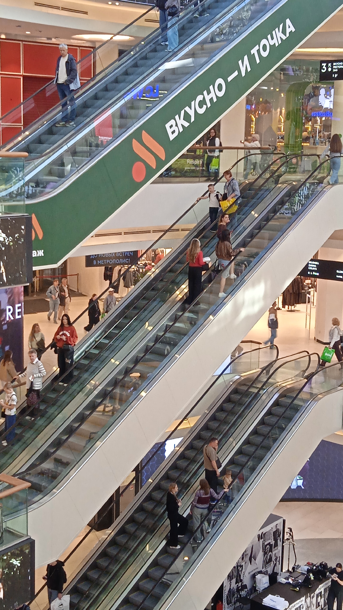 The image depicts the interior of a multi-level shopping mall, likely in Khimki, Russia, as evidenced by the Russian text visible on signage. Escalators are a prominent feature, with several people ascending and descending them. A large green banner with orange circular graphics and white text reading "ВКУСНО—И ТОЧКА" (which translates to "Tasty—and that's it") is prominently displayed across one of the escalators. Some people are casually dressed, while others appear to be shoppers carrying bags. The environment is brightly lit, typical of a modern retail space. The perspective is from a high vantage point, looking down at the escalators and the shoppers.