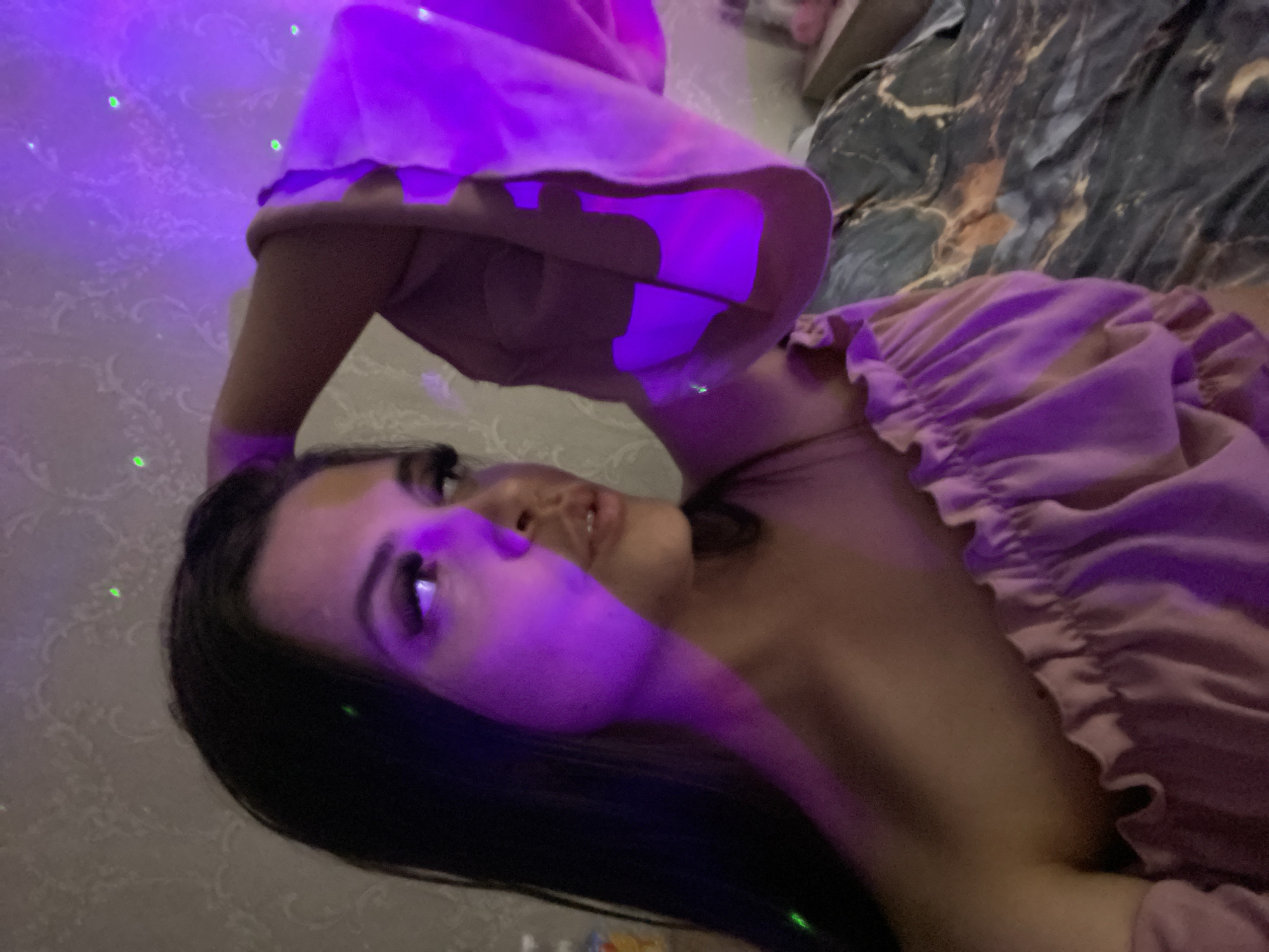 A woman with long, dark hair is captured in a medium close-up shot, looking to her right with her left arm raised behind her head. She is wearing a pink, off-the-shoulder top with ruffled detailing. The scene is illuminated by purple and green lighting, casting colored reflections on her skin and the background. The wallpaper behind her has a textured, damask pattern. To the right of the woman, a bed with dark, marbled bedding is visible. The image appears to be taken indoors, possibly in a bedroom, with decorative lights creating a vibrant atmosphere. There is no discernible information within the image to suggest a specific location in Chaikovsky, Russia.