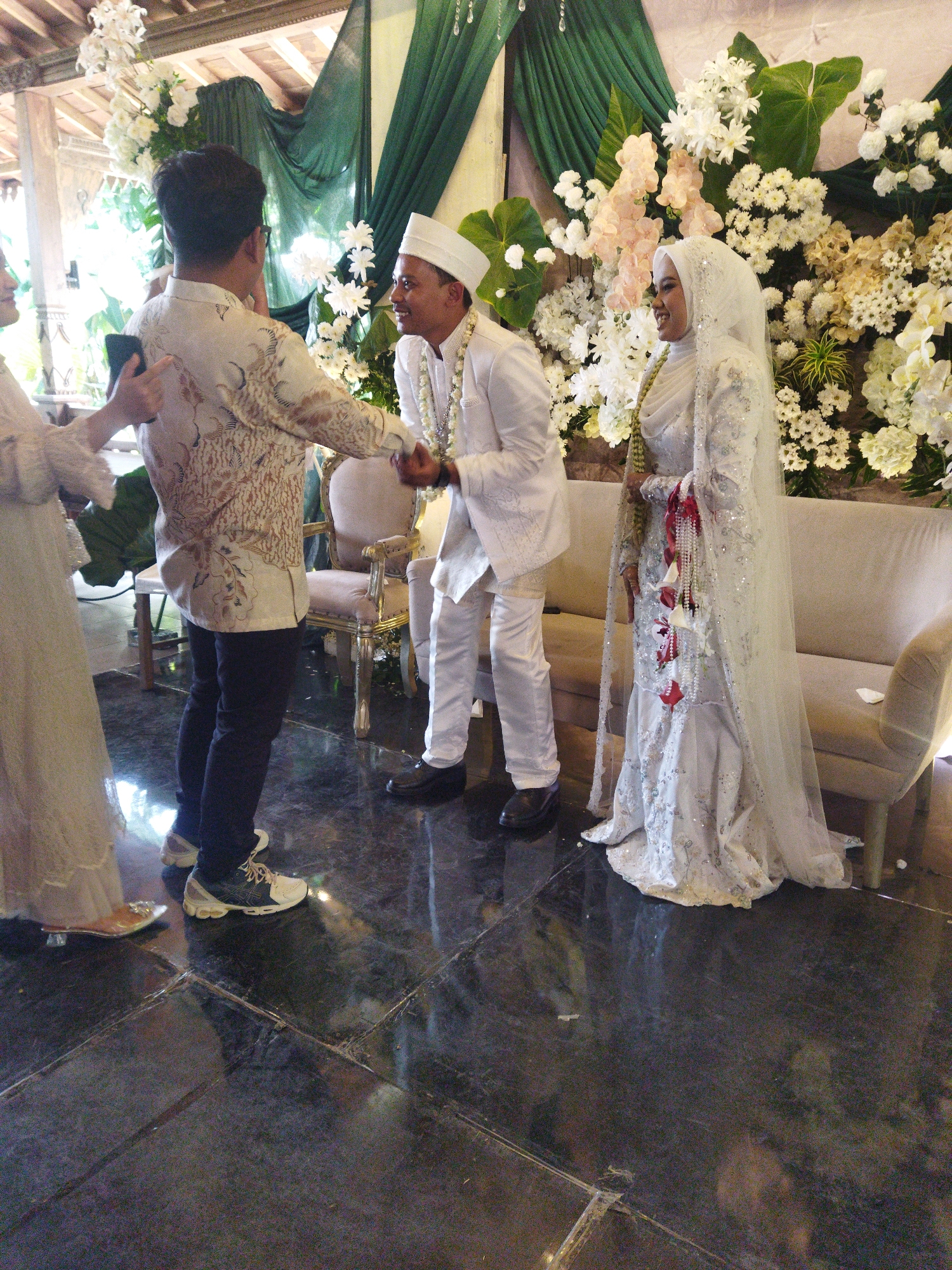 This image depicts a wedding ceremony at the Gedung SCJ (Semarang Convention Center) in Semarang, Indonesia. The foreground is dominated by a polished black floor, likely marble, reflecting the surroundings. In the mid-ground, a couple, the bride and groom, are engaged in a handshake. The groom, wearing a white peci (cap) and a matching white outfit adorned with intricate embroidery, is smiling. The bride is dressed in a traditional white wedding gown with a hijab and veil, also embellished with glitter and sequins, and a decorative beaded necklace. To the left of the groom, another man in a patterned cream-colored shirt and dark pants is seen shaking hands with him, holding a mobile phone in his left hand. Further to the left, a woman in a long cream-colored dress is partially visible. The background is a richly decorated setting with an abundance of white and pink flowers, green drapery, and a plush cream-colored sofa. The overall atmosphere suggests a formal and celebratory event, likely indoors given the lighting and architectural elements.