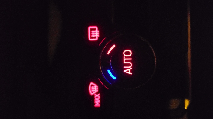 The image displays an illuminated vehicle climate control panel in a dark environment, purportedly located in Magaluf, Spain. A central circular dial features the word "AUTO" in red illumination. An arc of blue light is visible on the upper-left perimeter of this dial, and an arc of red light is on the upper-right. Above and to the left of the central dial, a red-illuminated button or indicator shows a windshield defog symbol with the text "MAX." Above and to the right, another red-illuminated button or indicator displays a rear defog symbol. The overall scene is dark, with only the control panel elements emitting light. A faint yellow light source is visible in the lower-left corner, and a subtle horizontal line of orange light is present along the bottom edge of the frame. No individuals or specific actions are depicted.