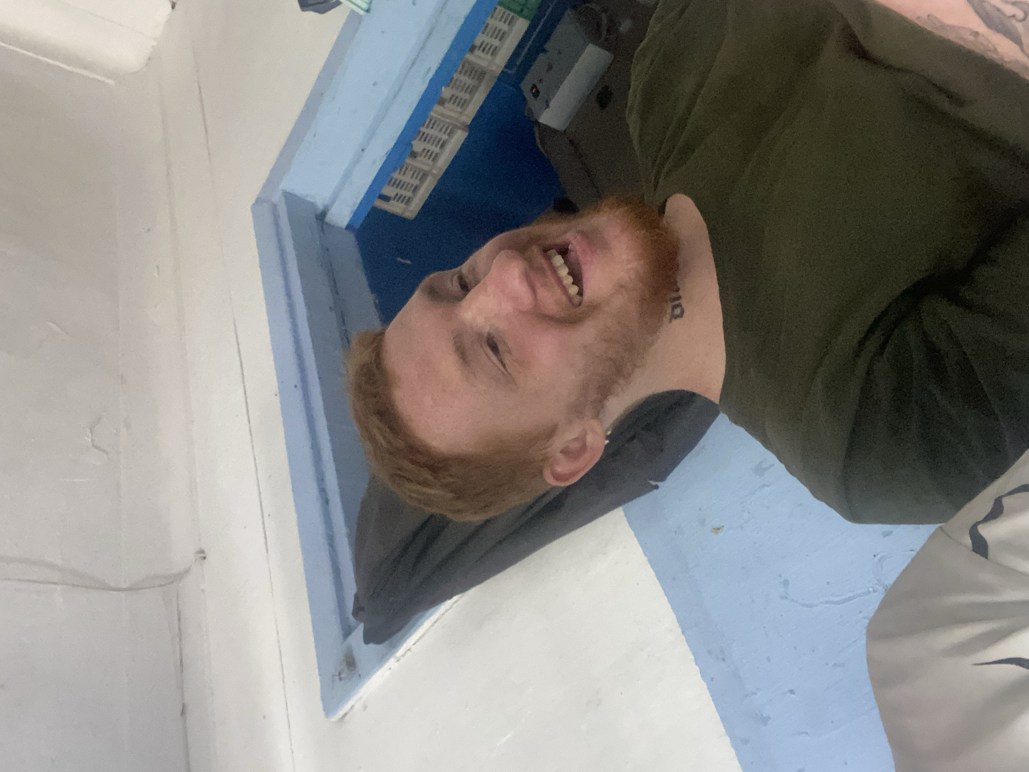A young Caucasian male with reddish-brown hair and a beard is lying down in a bunk bed. He is wearing a dark green t-shirt and has a tattoo on his neck. The bunk bed is painted blue and white, and there are some white crates on a shelf above him. The walls and ceiling are white. The scene appears to be inside a vehicle, possibly a train or a bus.