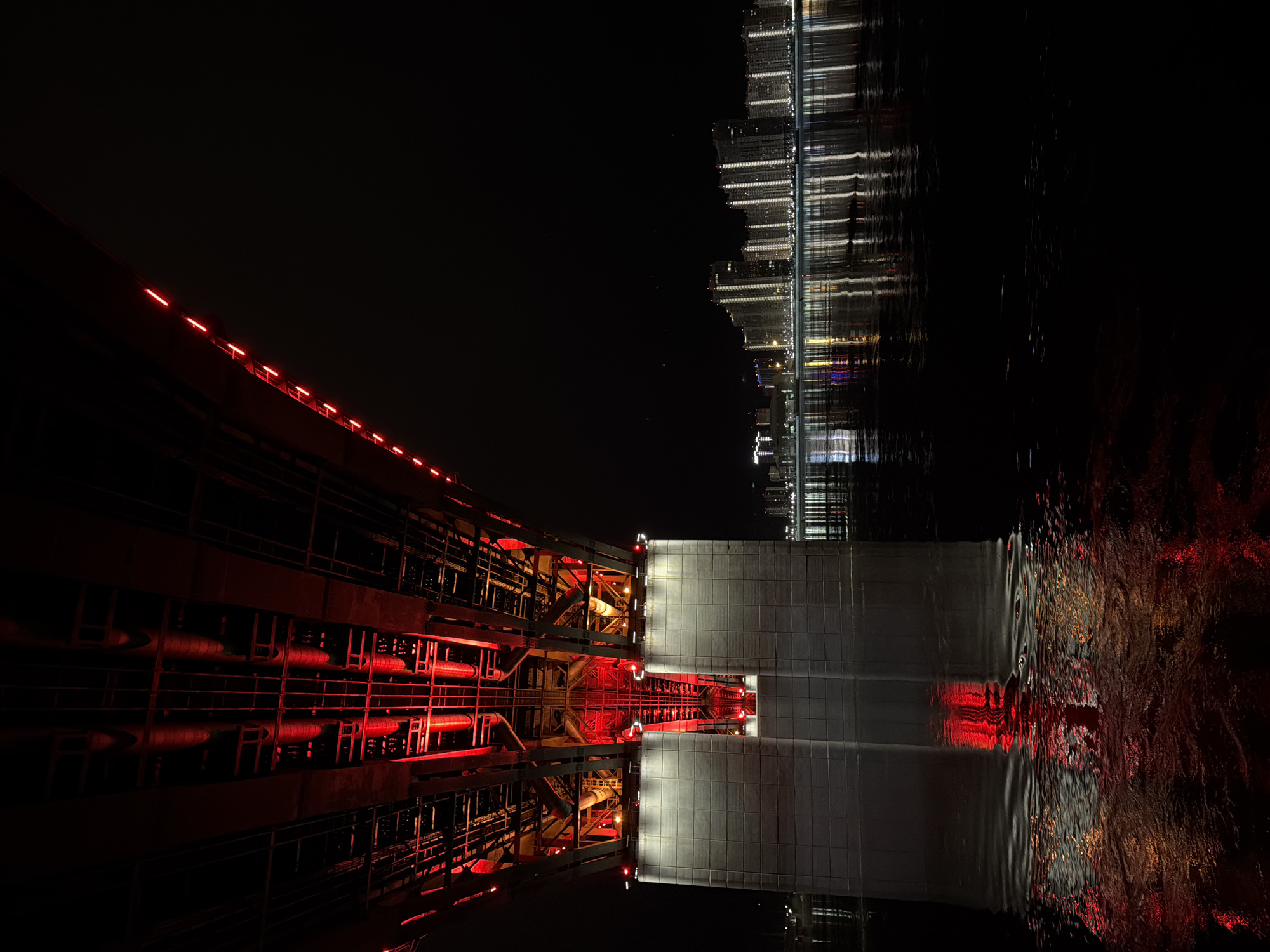 The image depicts a night view of the Vinogradovsky Bridge in Krasnoyarsk, Russia, reflected in the Yenisey River. The bridge's structure is visible, with red lighting illuminating its metal beams and supports. The river's surface shows a distorted reflection of the bridge and the city lights in the background. The right side of the image shows a reflection of city buildings with white lights.