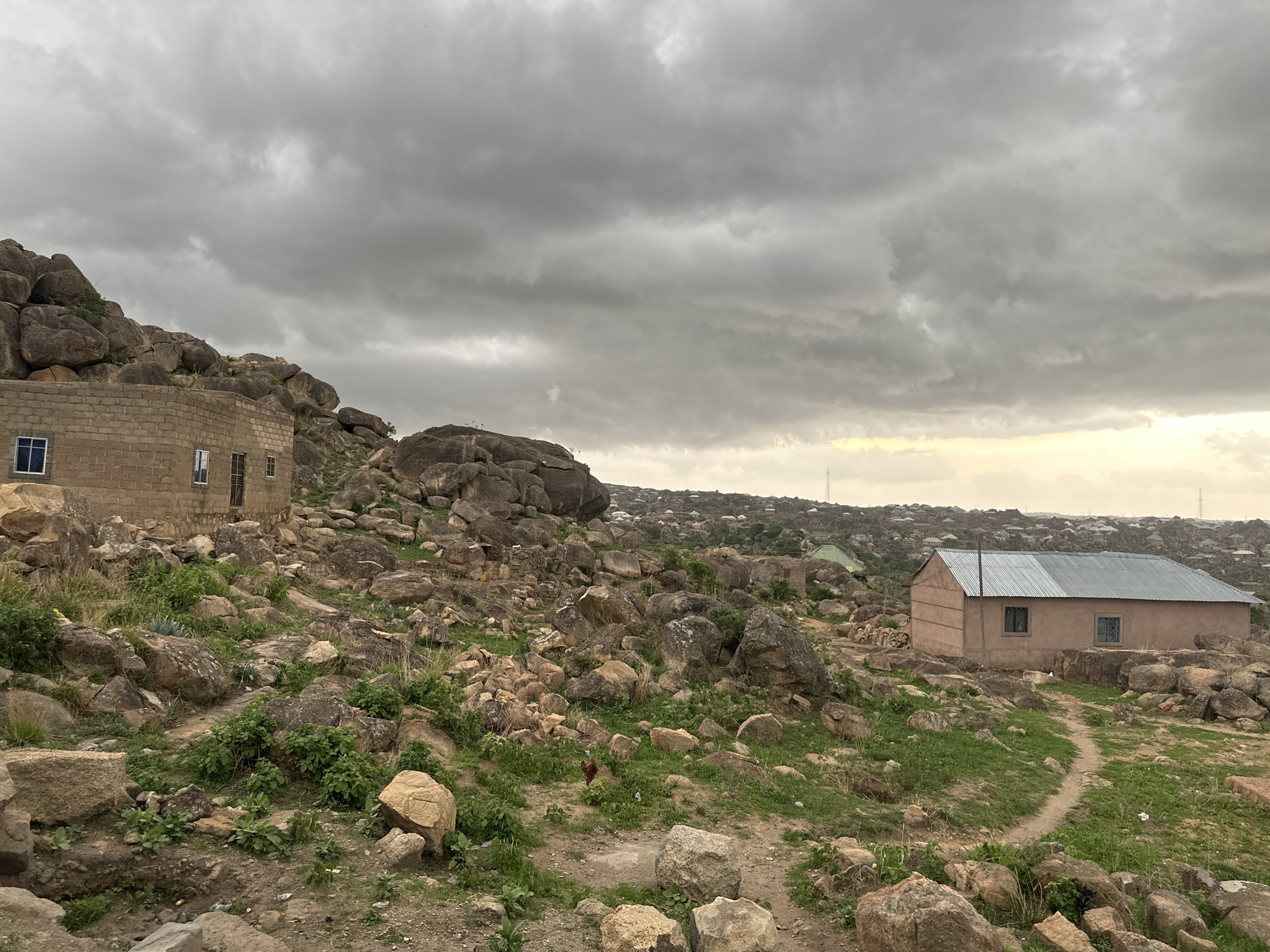 The image shows a rocky, hilly landscape under a cloudy sky. Two buildings are present. The building on the left is constructed of tan-colored blocks, while the building on the right is tan with a corrugated metal roof. Rocks and sparse vegetation cover the ground. A dirt path is visible leading towards the building on the right. In the background, a denser collection of buildings indicates an urban area. A single chicken is visible on the ground in the foreground. The location is recognizable as a rocky, elevated area in Jos, Nigeria based on the buildings and overall terrain.