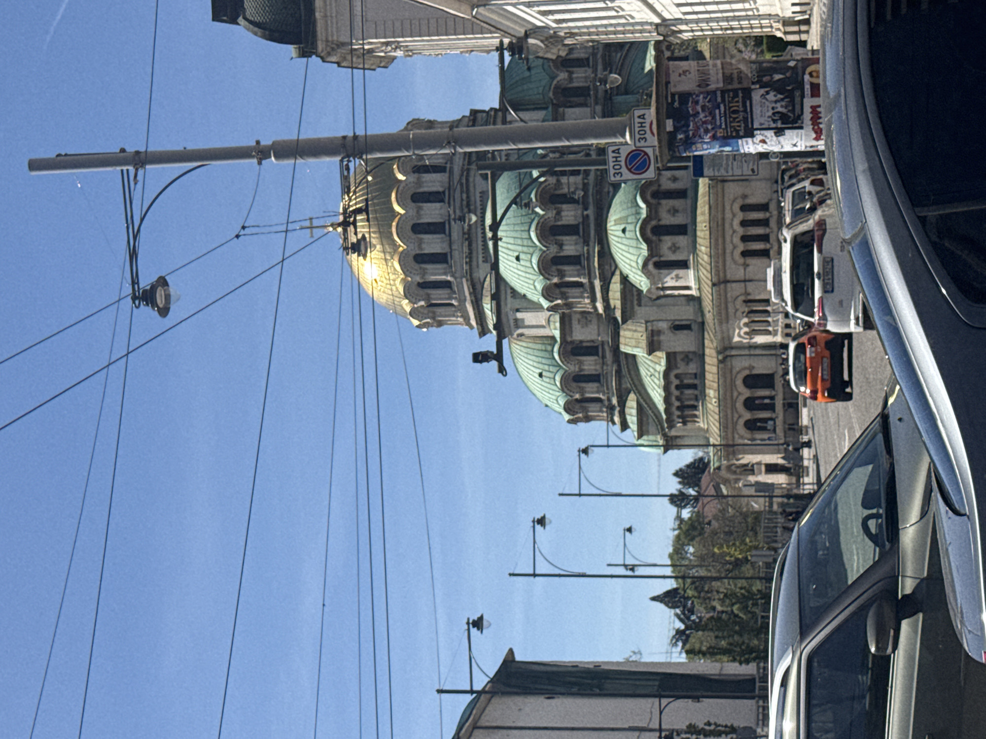 The image captures an urban street scene in Sofia, Bulgaria, dominated by the majestic Alexander Nevsky Cathedral on a bright, sunny day. The impressive building features a prominent golden central dome reflecting sunlight, surrounded by several smaller teal-green domes. Its facade is light-colored stone with intricate architectural details, including arched windows and decorative elements.
The scene is oriented vertically, showing a wide, clear blue sky. In the foreground, the side of a dark-colored car is partially visible on the right, suggesting it is parked along the street. Further down the street, a white car and an orange car are parked or moving.
Utility poles and streetlights line the street, with numerous overhead wires crisscrossing the sky, indicating a trolleybus or tram network. A tall, gray traffic pole stands prominently in front of the cathedral, displaying two traffic signs. The signs feature Cyrillic text "3OHA" (ZONA, meaning "Zone"). One is a blue square sign with "30HA" written, typically marking the beginning of a zone. The other is a red circular sign with a diagonal white bar over "30HA", which generally signifies the end of a zone (likely a 30 km/h speed limit zone, given the "30"). In the background, some posters with Cyrillic text are visible on a utility box or wall, though the text is illegible. Lush green trees can be seen behind the buildings. The bright sunlight suggests it is daytime, possibly morning or early afternoon, with clear weather.