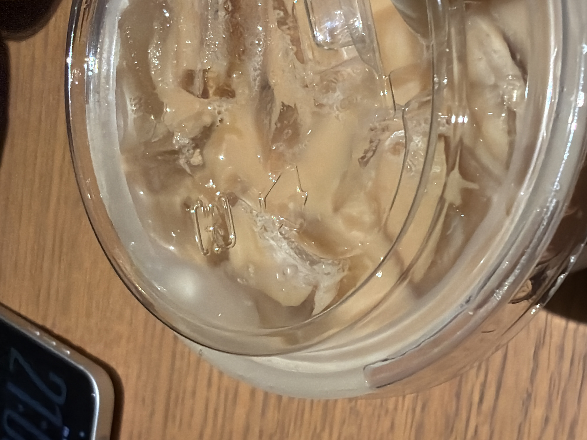 An overhead, close-up shot shows an iced coffee drink in a clear plastic cup with a lid. The drink is light brown and contains ice cubes. A smartphone with a black screen displaying a time of "21:12" is visible in the upper left corner of the image, resting on a wooden surface. The wooden surface appears to be a table. There is no indication of the city of Đà Nẵng, Vietnam, in this image.