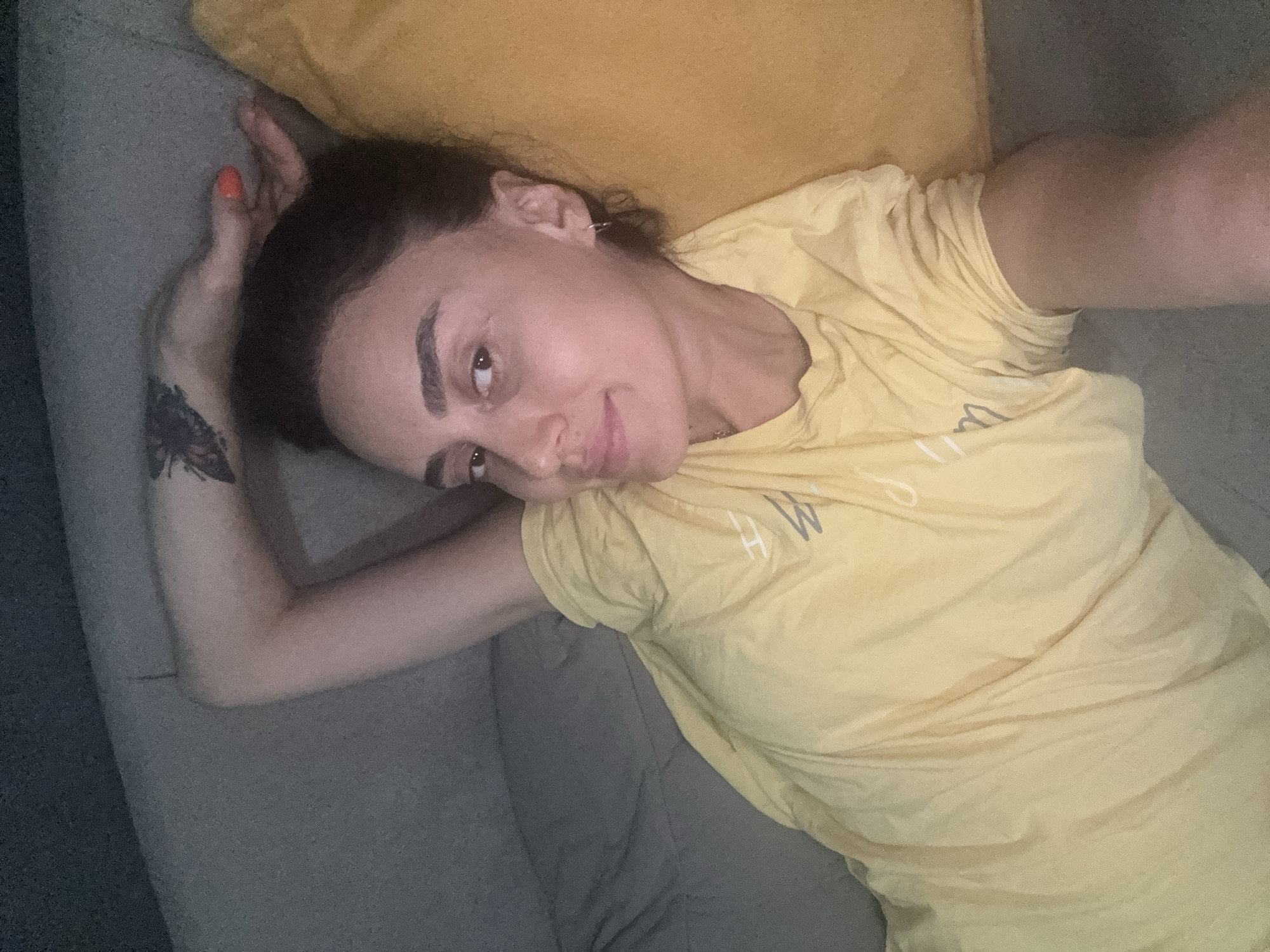 In Tbilisi, Georgia, a woman with dark hair and a butterfly tattoo on her left arm lies on a sofa, resting her head on a yellow pillow. She is wearing a yellow t-shirt. Her left arm is bent, with her hand near her head. The sofa is a neutral grey.