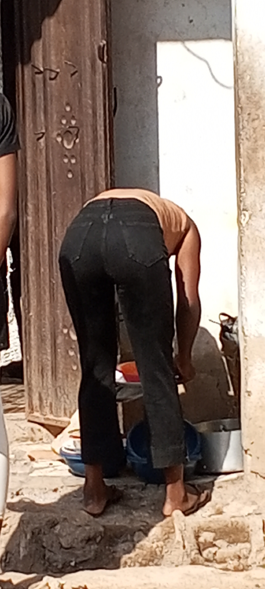 A person wearing dark jeans and a light brown top is bending over, appearing to be interacting with objects near a wall and what appears to be a bucket. The person is wearing sandals or flip-flops. Another person is partially visible to the left, wearing a dark shirt. The background includes a detailed wooden door and a white wall. The location is indicated as Sabon Tasha, Nigeria.