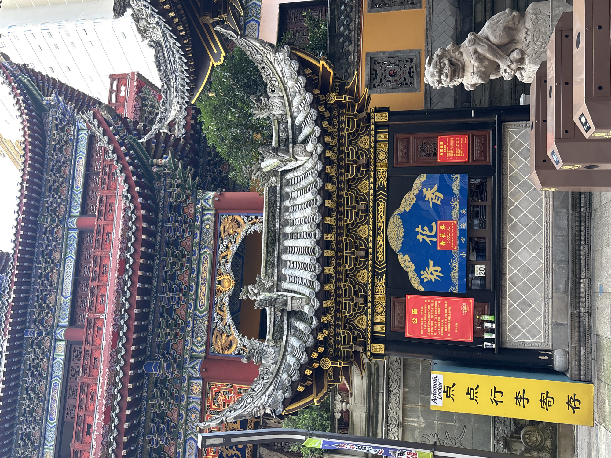 The image depicts the entrance of a traditional Chinese temple in Yuzhong District, China. The temple features ornate architecture with multi-tiered, curved roofs adorned with intricate colorful carvings and dragons. In the foreground, a stone lion statue sits to the right of the entrance, guarding the premises. To the left of the entrance is a bright yellow sign with Chinese characters and the words "Automatic Locker." A red announcement poster is affixed to the wooden entrance door. Behind the entrance, a blue banner with golden Chinese characters is displayed. A modern building is visible in the background, contrasting with the traditional temple architecture. There are no people visible in the frame. The environment appears to be an outdoor public space.
