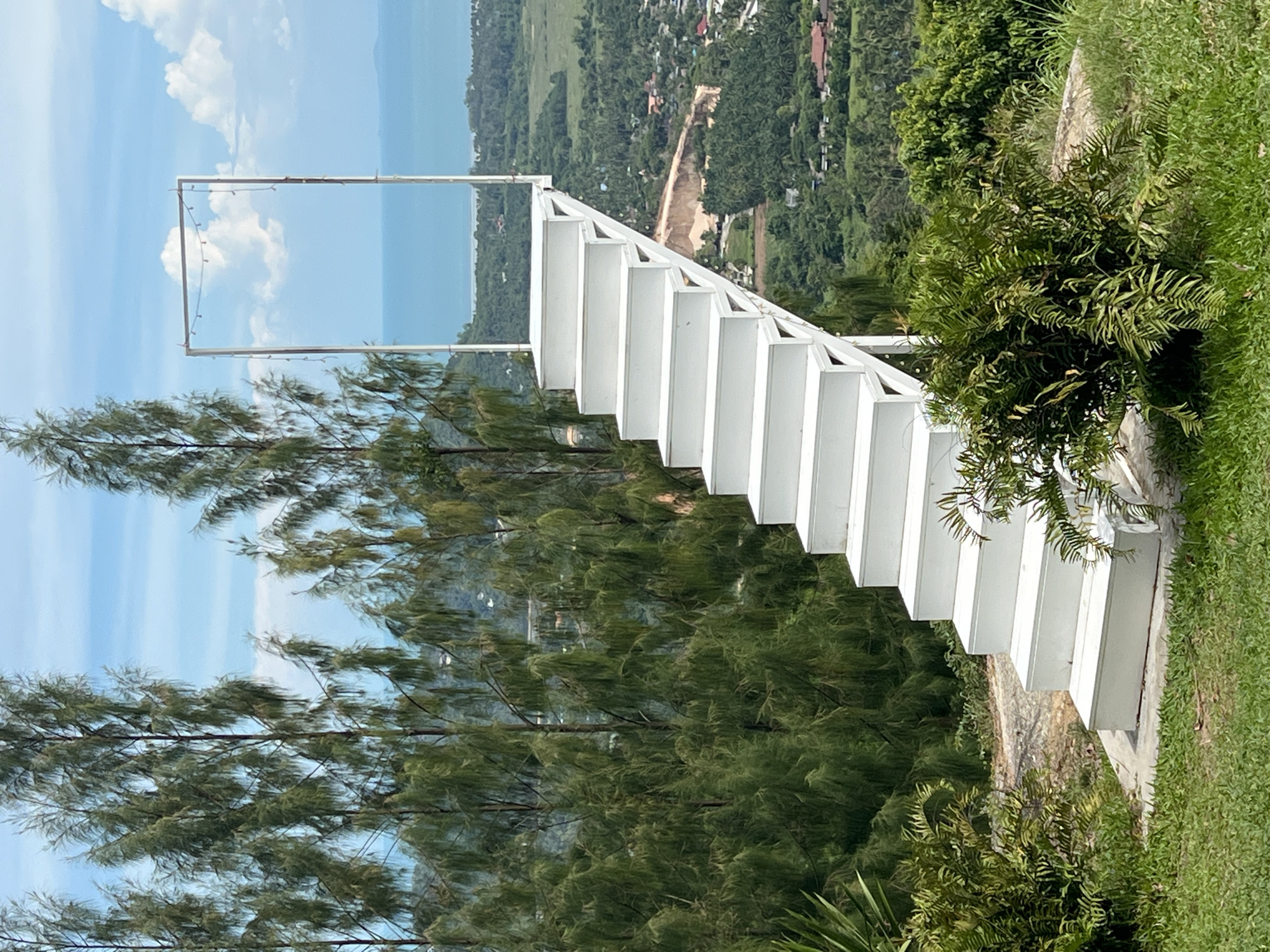 A bright white staircase ascends from a grassy area towards a rectangular metal frame with lights. The staircase is built of wood and has many steps. The metal frame is situated at the top of the stairs and appears to be a doorway opening onto a panoramic view of a lush green valley with scattered buildings and a body of water in the distance under a blue sky with white clouds. Tall, slender trees with feathery green foliage surround the staircase. The scene is located in Baan Thurian, Thailand, and is a scenic overlook or viewpoint. There are no people or animals visible in the image.