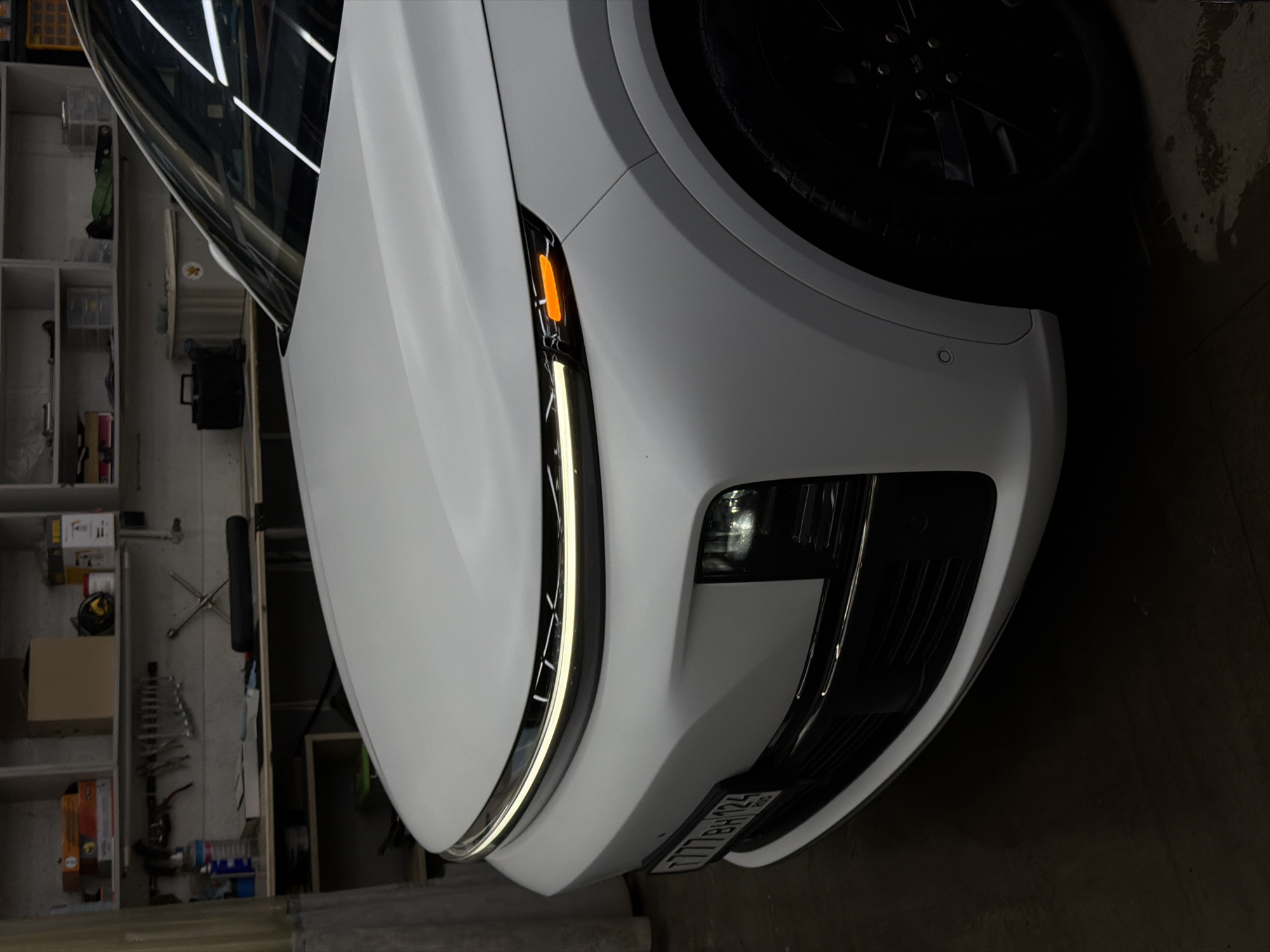 A white car, possibly a sedan or SUV, is shown parked indoors, likely in a garage or workshop. The image is taken from a low angle, focusing on the front-right quarter of the vehicle, rotated clockwise. The car features modern design elements, including a sleek white body, a black grille, and distinctive LED daytime running lights that form a continuous strip along the top edge of the headlight assembly. An amber side marker light is illuminated on the front fender, and a lower fog light or auxiliary lamp is also visible. The front tire and black multi-spoke wheel are partially seen in the upper right.
The background reveals a utilitarian setting with shelving units filled with various items like boxes, tools, and bottles. Reflections of overhead fluorescent lights are visible on the car's windshield and the glossy white paintwork, indicating an indoor, artificially lit environment.
A license plate is visible on the front bumper, displaying the text "777 BH 124". The "124" region code on the license plate corresponds to Krasnoyarsk Krai, Russia, aligning with the provided location context of Krasnoyarsk. The car appears clean and well-maintained. No specific activity is depicted beyond the car being stationary.