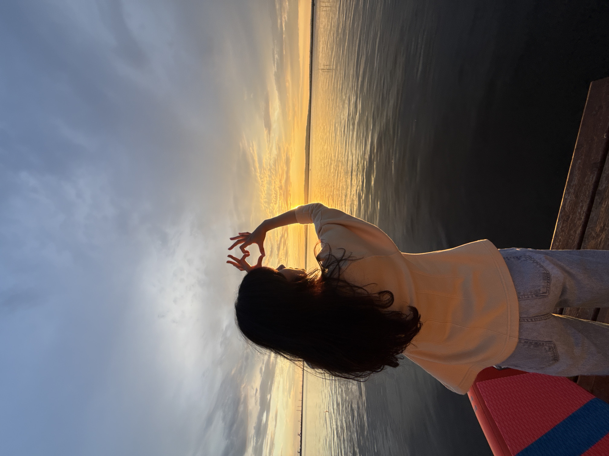 A person with long, dark hair stands on a wooden dock, facing away from the camera. They are wearing a light-colored jacket and light-wash jeans. Their hands are held up, forming a heart shape against the sky. The background is a body of water, likely the Kama River, with a vibrant sunset casting an orange glow on the horizon. The sky is partly cloudy, with shades of blue, grey, and orange. The person appears to be in Chaikovsky, Russia, given the context. The scene conveys a sense of peace and appreciation for nature.