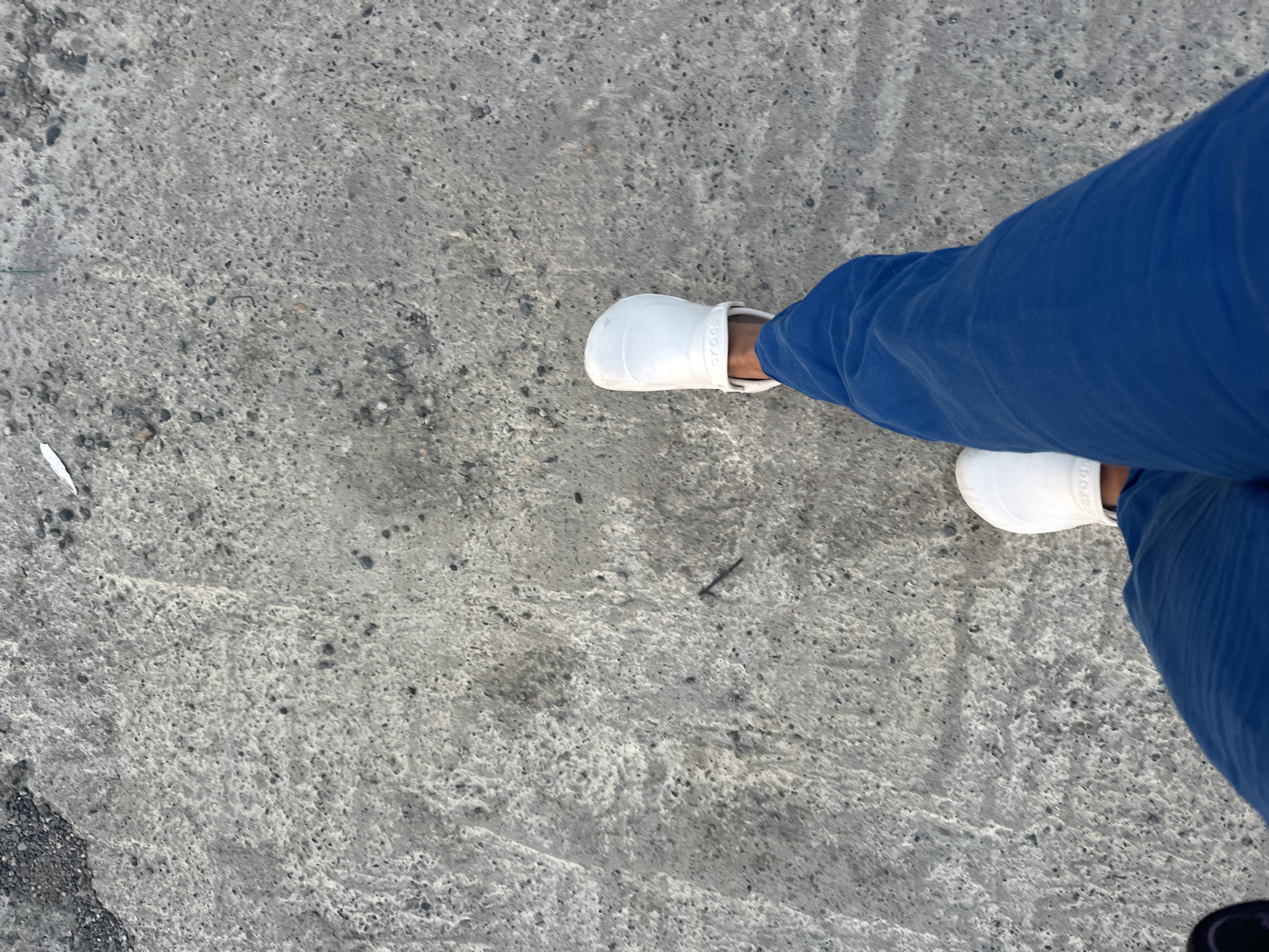An overhead shot shows a pair of feet wearing white Crocs and blue pants walking on a gray concrete surface. The concrete is rough and textured, with small pebbles and darker patches visible. The perspective is from the person's viewpoint, looking down at their own legs and feet as they move forward. The background is entirely the concrete ground. The specific location in Razdolnoye, Russia, cannot be determined from the image alone.