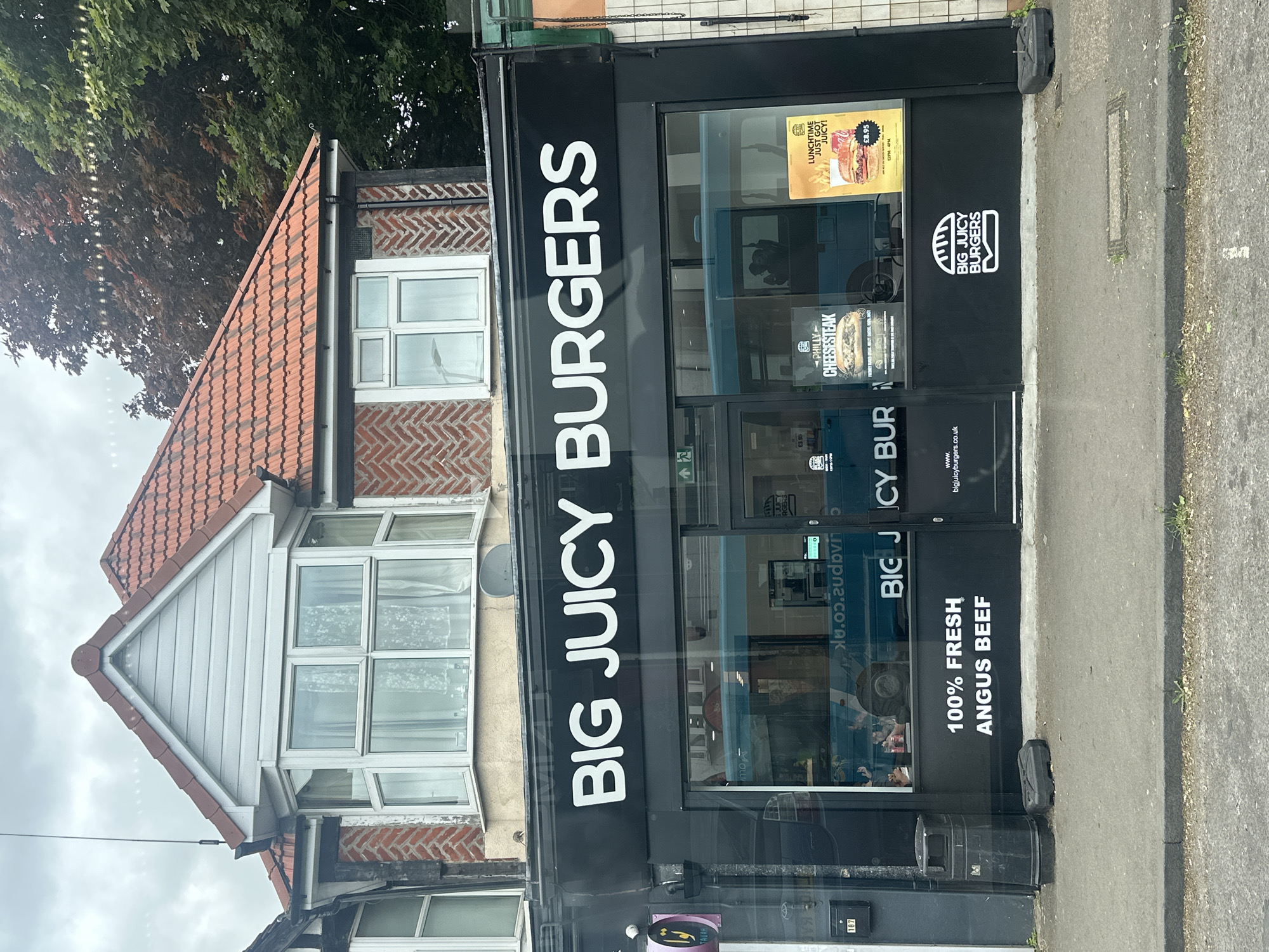 The image shows the storefront of "Big Juicy Burgers" in St Albans, United Kingdom. The store has a black facade with white lettering. The name "BIG JUICY BURGERS" is prominently displayed above the entrance. A menu board is visible in the window, advertising "LUNCHTIME JUST GOT JUICY!" for £8.95, and "PHILLY CHEESESTEAK" for £3.95. The restaurant's logo is displayed on the front. The store's address, "187", is visible on the building's exterior. A residential building is adjacent to the restaurant, with a red tiled roof and a bay window. A trash receptacle is adjacent to the main building.