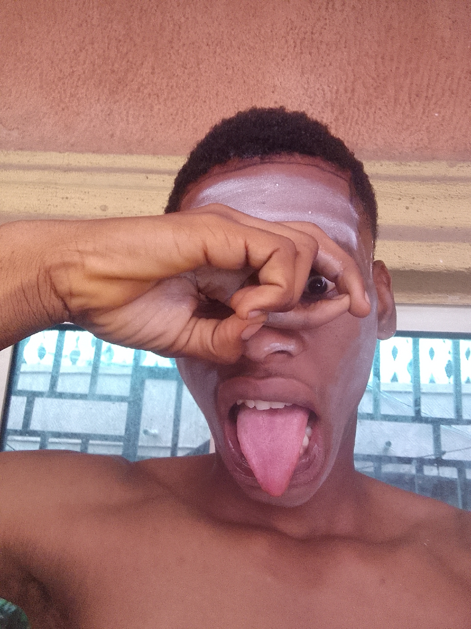A close-up portrait of a shirtless, dark-skinned young man. He is sticking out his tongue and forming an "OK" symbol with his right hand, peering through the opening. A white substance is smeared across his forehead. In the background are window frames, likely in Nkpor, Nigeria.