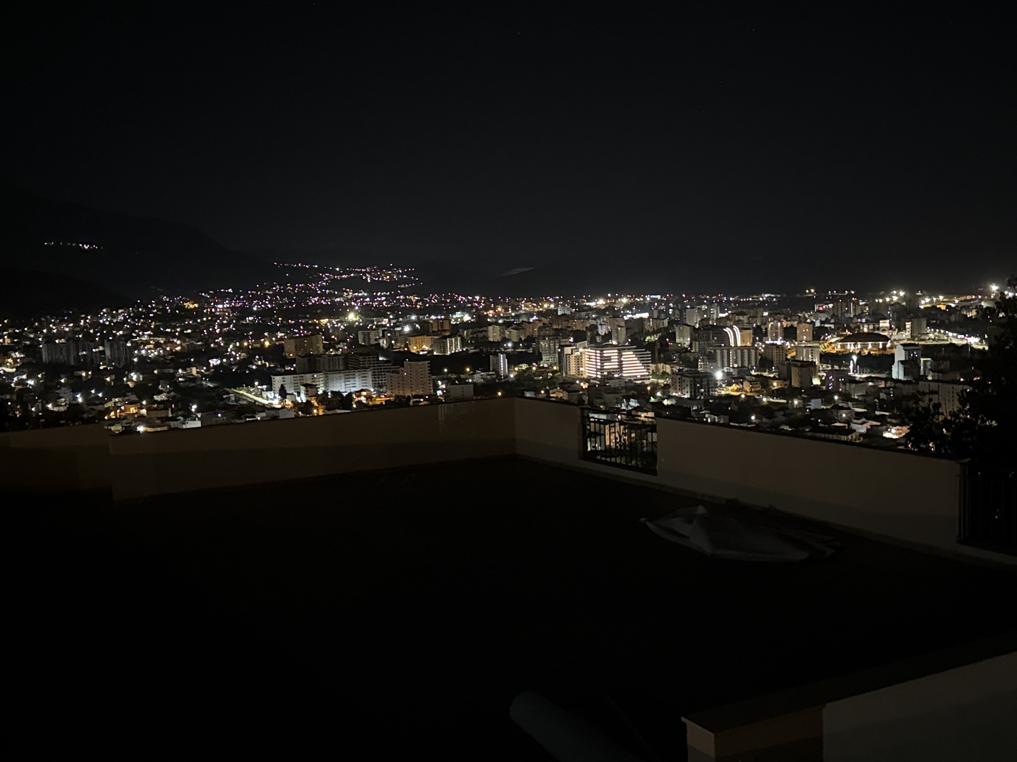 A low-angle, wide shot captures a cityscape at night from a high vantage point. The darkness of the night sky is contrasted by the numerous lights of the city stretching across the horizon. A mountain range is dimly visible in the background, punctuated by sparse lights indicating higher elevations.
The foreground is dominated by a dark, flat surface, presumably a rooftop or balcony, enclosed by a low concrete wall. The wall is interrupted by sections of black metal railing. On the dark surface, a light-colored object is partially visible, appearing like a folded textile or covering. A portion of what appears to be furniture can be seen along the bottom of the view.
The overall atmosphere is quiet and still. The city is Šušanj, Montenegro.