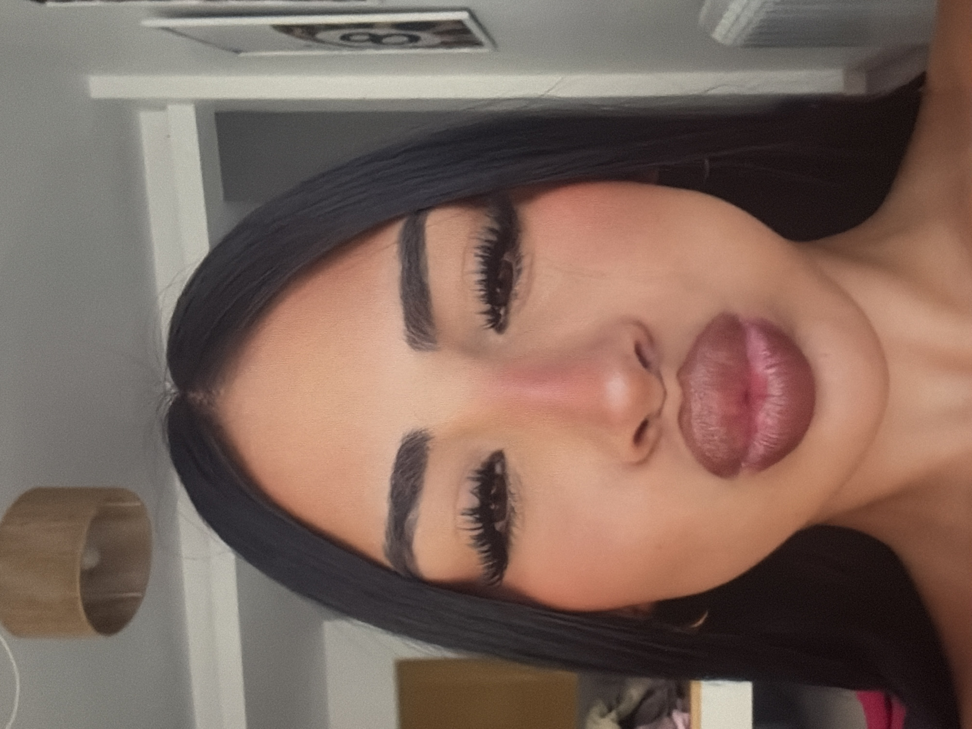 The image is a close-up, front-facing selfie of a woman with dark hair, dark eyebrows, and long eyelashes. She is wearing makeup, including blush and lipstick, and her lips are pursed. She is wearing earrings. The background is blurred but appears to be an indoor setting with a ceiling light fixture, a shelf with some items, and a framed picture on the wall. The lighting is soft and diffused.
