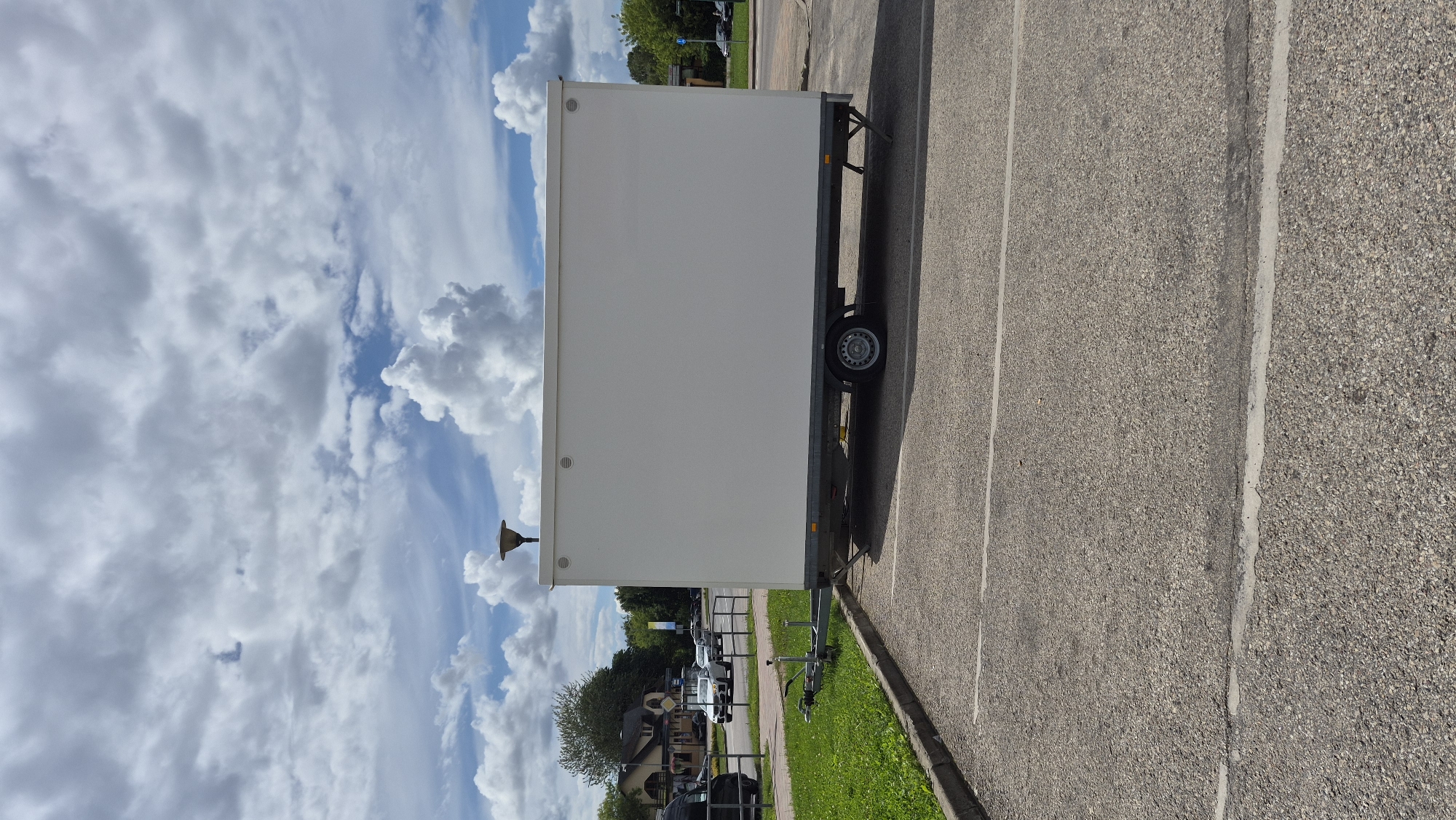 A white trailer, possibly a portable restroom unit, is parked in a paved parking lot next to a grassy area and a road. The trailer is rectangular with a flat roof and a single wheel on each side. A hitch is visible at the front of the trailer. A street lamp is mounted on top of the trailer. In the background, there is a road with several cars, a building with a red roof, and trees. The sky is partly cloudy. The scene appears to be located in Jēkabpils, Latvia.