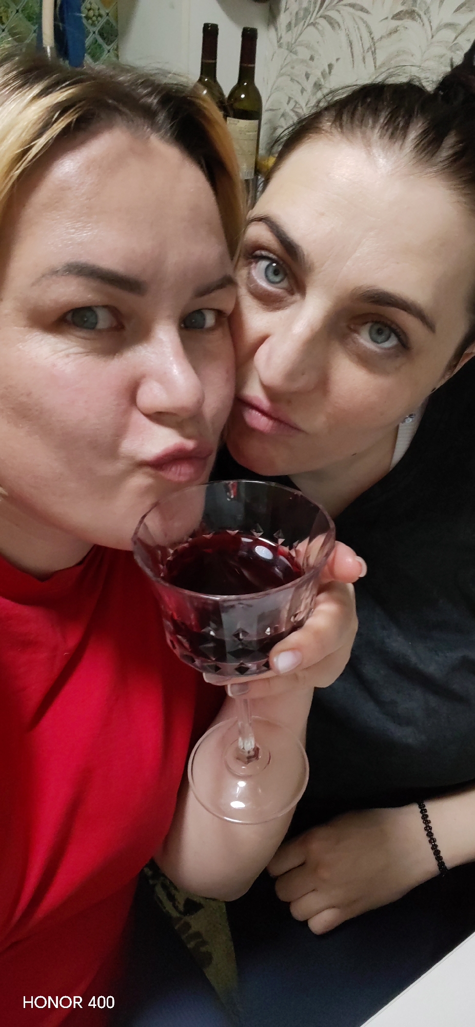 The image displays a close-up, chest-up view of two adult females indoors. The female on the left has blonde hair with dark roots, light blue eyes, and fair skin, wearing a red garment. She presents a puckered-lip expression and looks directly at the camera. The female on the right has dark hair styled in an updo, light blue eyes, and fair skin, wearing a dark-colored top with a white strap detail visible on her right shoulder and a small, light-colored earring. She has a black beaded bracelet on her left wrist. She holds a clear, faceted stemmed glass containing a dark red liquid with her right hand and also looks at the camera. The two individuals are positioned tightly together. The background reveals an indoor setting with light-colored, patterned wallpaper behind them, and two partially visible dark green wine bottles are present on a surface. A section of a wall with light blue or green patterned tiles is visible to the far left. The specific city location of Noviy, Russia, cannot be determined from the visual information provided. The image includes an "HONOR 400" watermark in the lower-left corner.