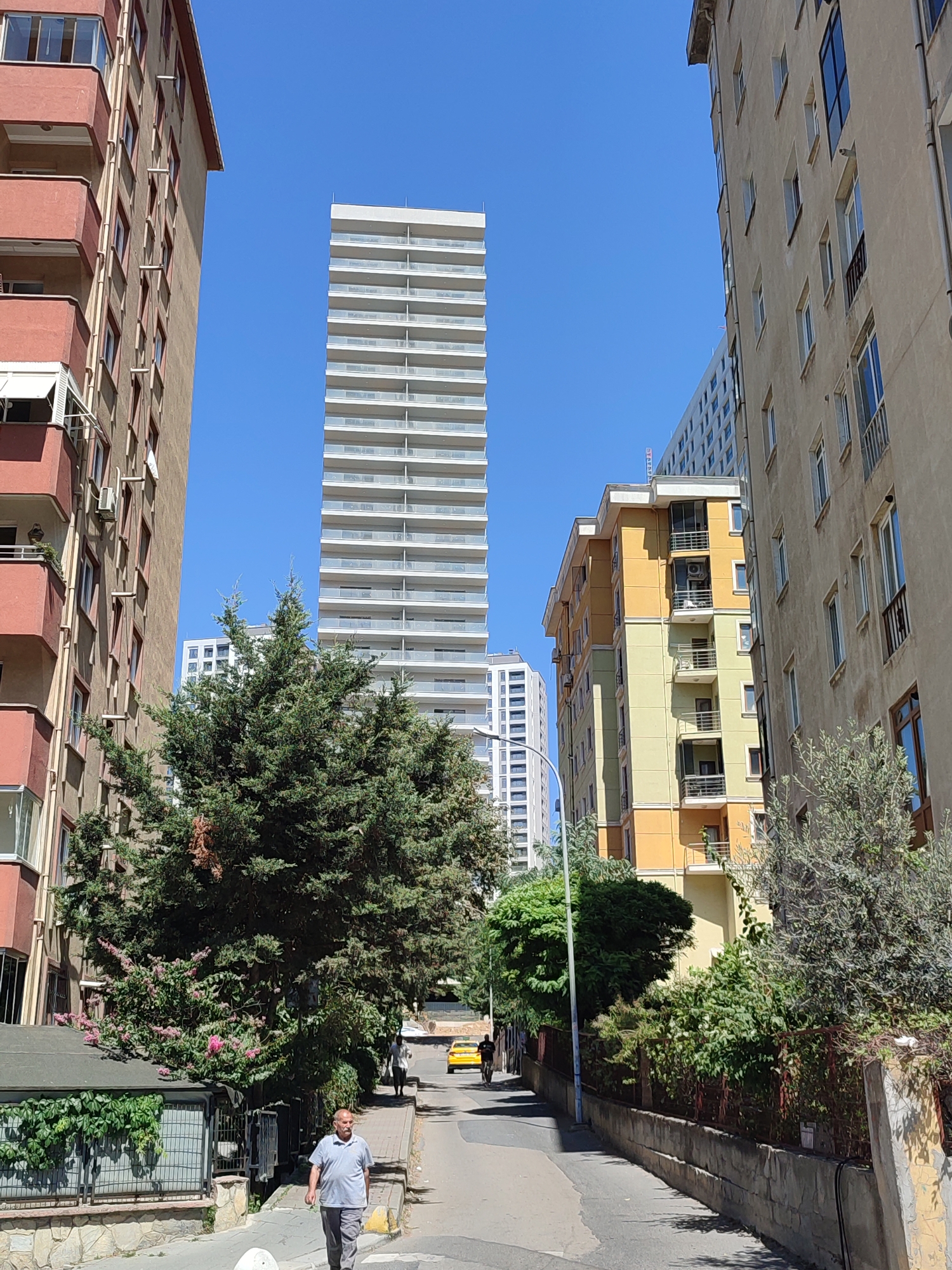 In Kadıköy, Turkey, a street scene unfolds under a clear blue sky. The foreground features a man walking away from the camera, dressed in a grey polo shirt and grey pants. To his left, a low, grey-roofed structure is covered in green ivy, with a grey fence in front. A large green tree with pink blossoms is to the right of this structure.
The mid-ground of the image shows a paved street with a yellow taxi driving away from the viewer. A tall, slender white skyscraper dominates the background, characterized by its many horizontal white balconies. To the left of this skyscraper, a reddish-brown apartment building with balconies is visible. To the right of the skyscraper, a smaller, multi-story building painted in alternating bands of yellow and pale green stands. Further to the right, a tan-colored apartment building with numerous windows and balconies occupies the edge of the frame. Lush green trees and foliage are scattered throughout the scene, softening the urban landscape.