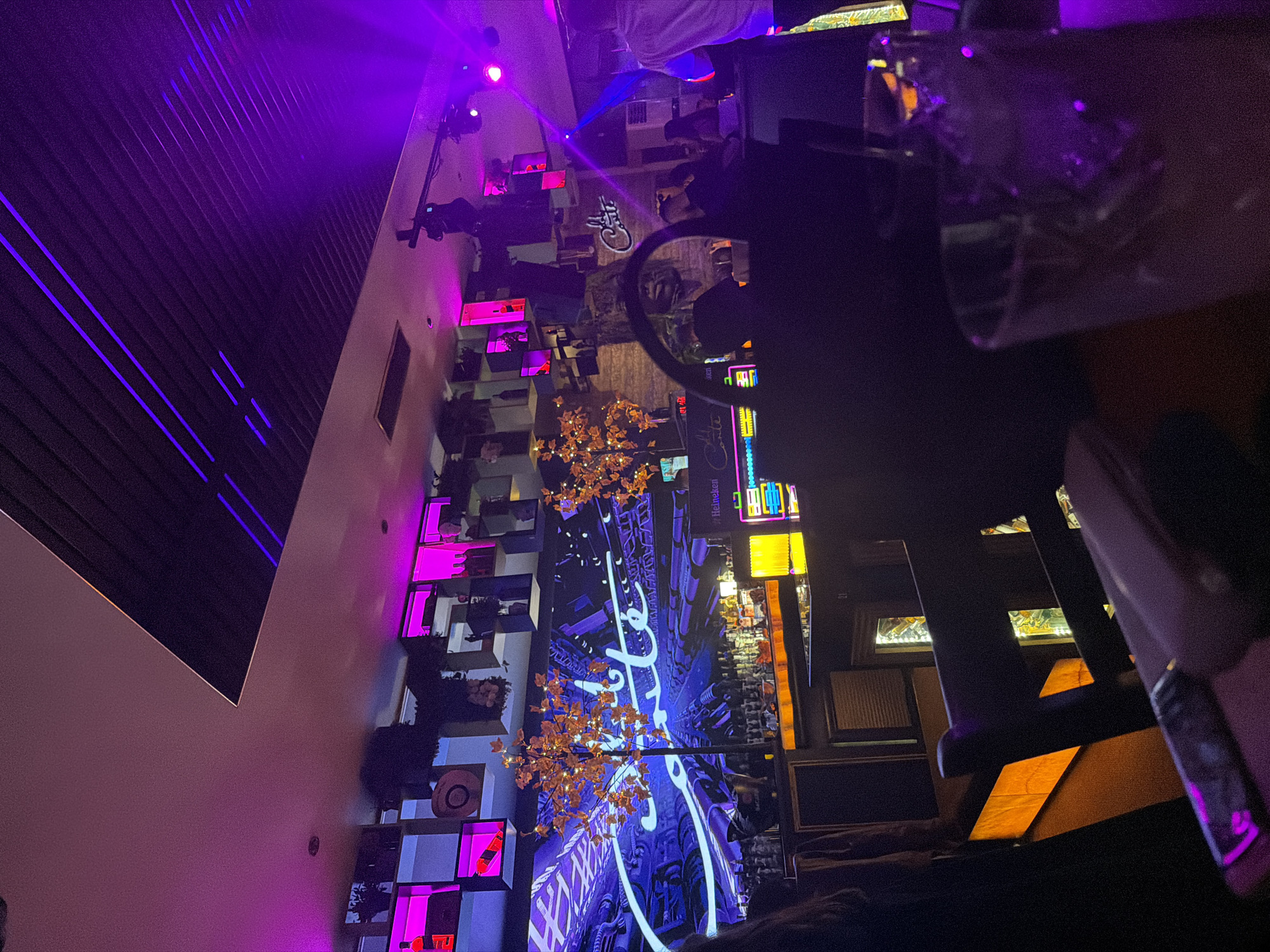 The image depicts the interior of a dimly lit bar, illuminated by purple and yellow ambient lighting. There are tables and chairs in the foreground, some obscured by foreground elements such as glasses. The walls are adorned with decorative elements including shelving with items on display, framed artwork, and neon signage. The space is populated with people, although many are blurred due to motion or depth of field. The bar is likely located in Sofia, Bulgaria, as indicated by the Heineken sign.