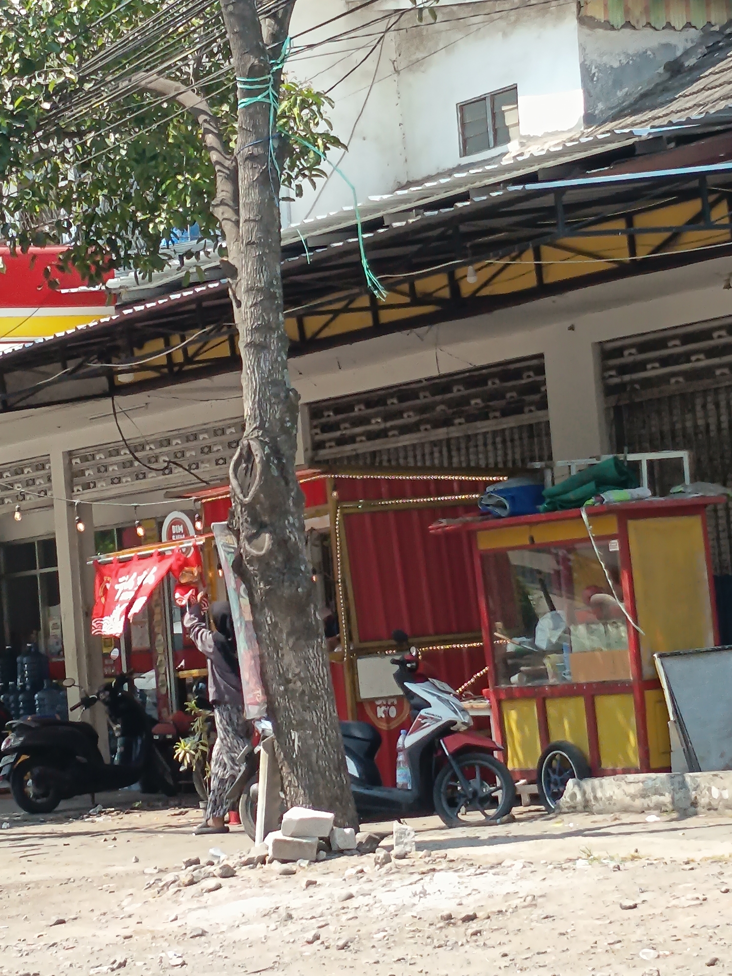 In the city of Kandangjati Kulon, Indonesia, a street scene unfolds with a prominent tree trunk in the foreground. The tree is cinched with light blue rope, suggesting it might be used for support or as a guide. Behind the tree, a red and yellow food stall with a glass display case is visible, its awning casting a shadow. Two motorbikes are parked on the dusty ground; a black scooter on the left and a red and white motorbike to the right. A person wearing a hijab and patterned trousers stands near the tree, facing the stall. The background reveals the facade of buildings with corrugated metal roofs and awnings, some with visible signs. Overhead, a tangle of electrical wires crisscrosses the sky. The ground is rough and unpaved, littered with small rocks and debris.