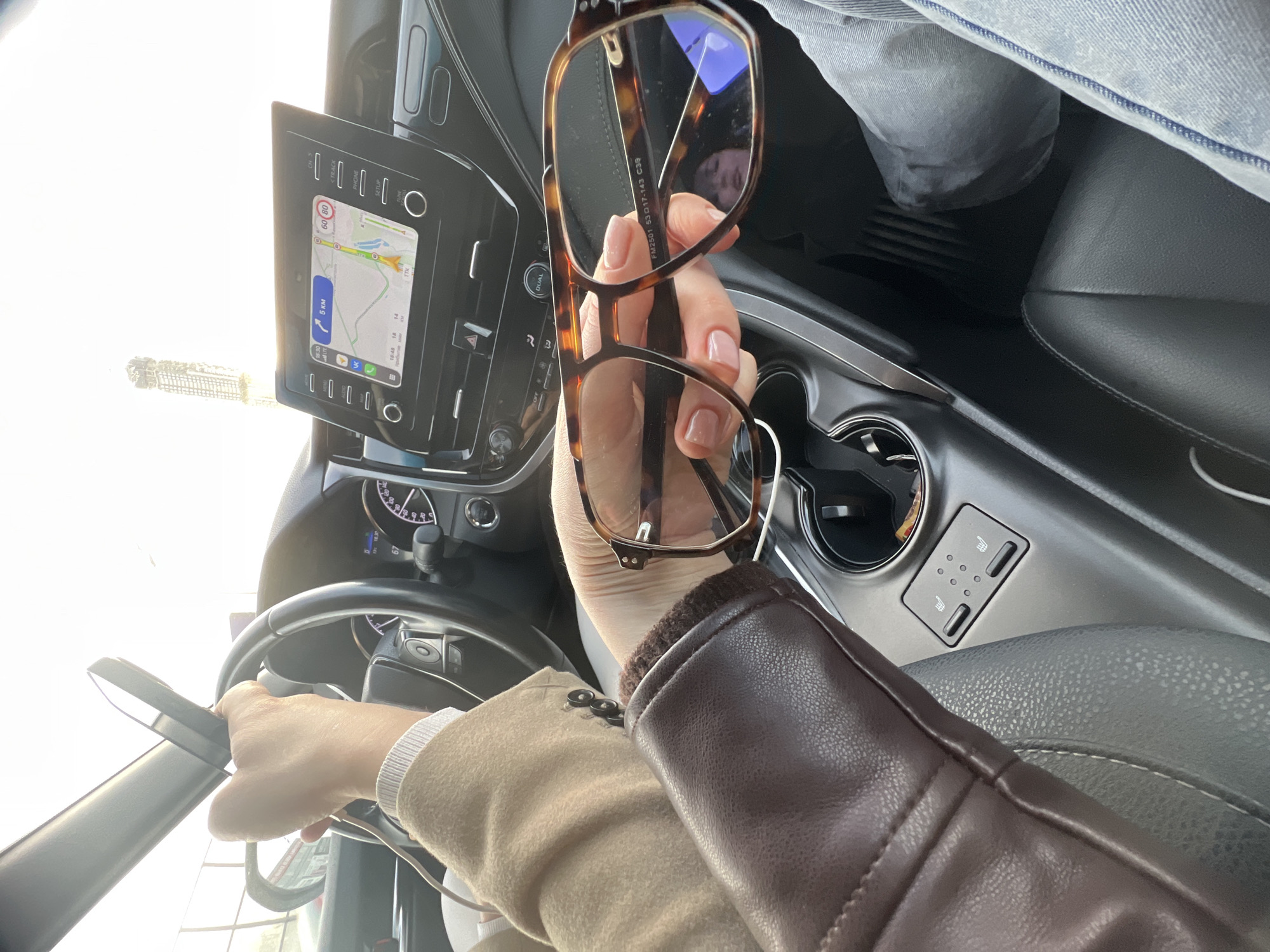 The image shows a person inside a car, likely in Moscow, Russia, based on the navigation display. The person is holding glasses in their right hand, which is adorned with a brown leather-sleeved jacket, while their left hand is on the steering wheel. The car's interior features a navigation screen showing a map with Russian text. The center console includes buttons and cupholders. The overall scene suggests the person is either stationary in the car or preparing to drive.