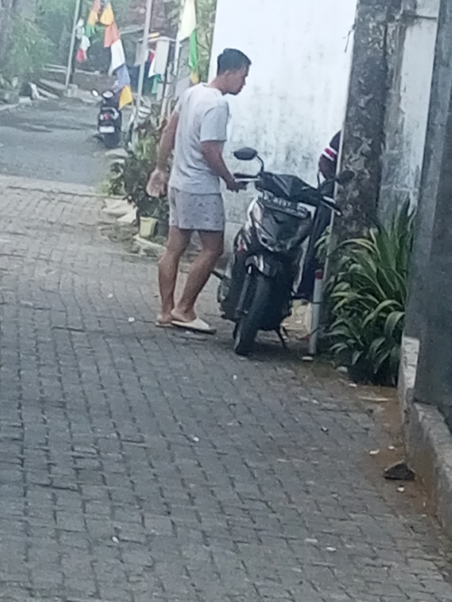 In Andongsili, Indonesia, a man wearing a light-colored t-shirt and patterned shorts stands next to a black motorcycle on a paved pathway. The man is facing away from the camera, and he appears to be holding a white object in his right hand. The motorcycle is parked on the right side of the pathway, with its front wheel turned to the left. In the background, there is a white wall and some greenery. The overall scene suggests a casual, everyday moment in a residential area.