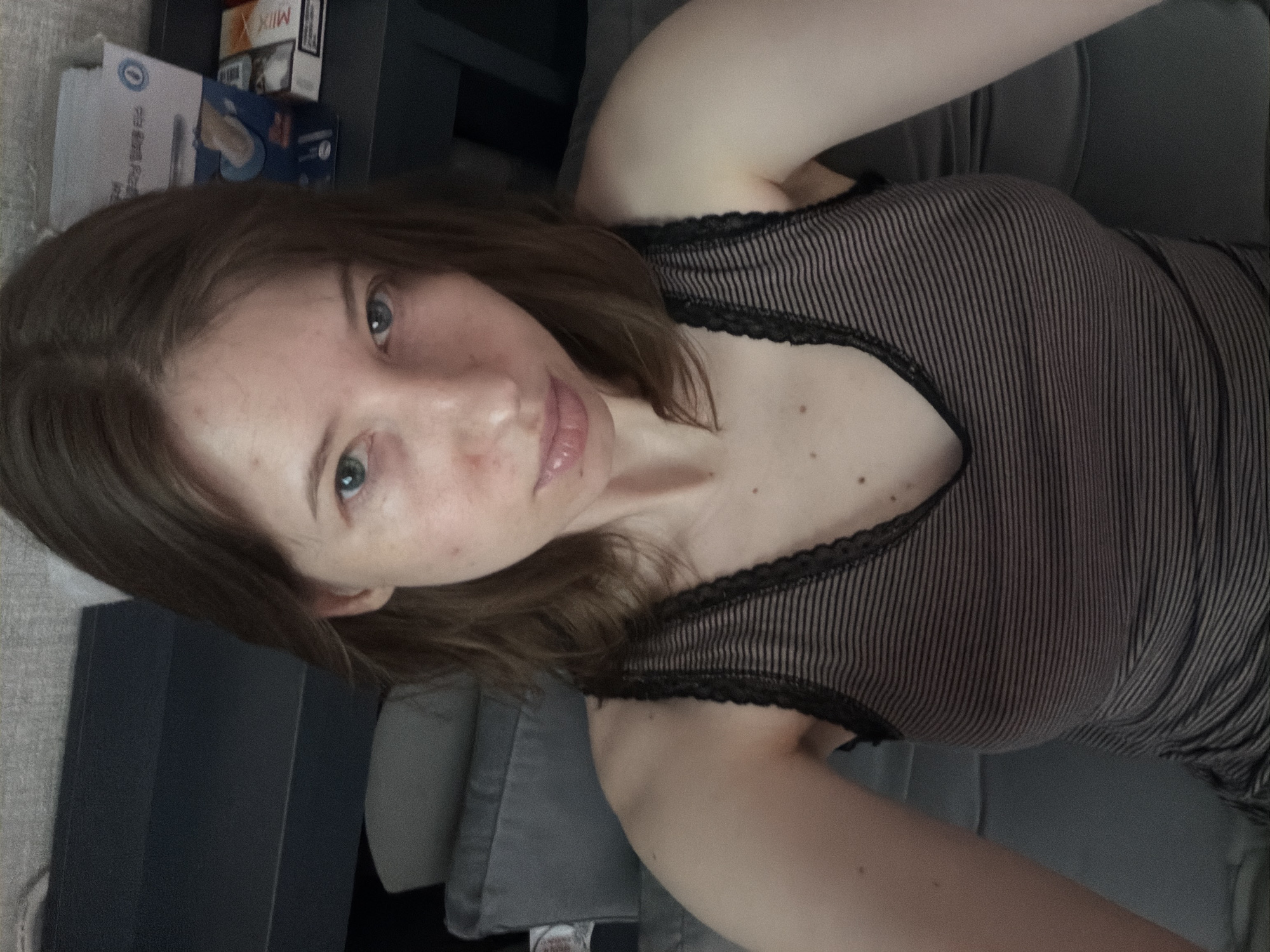A young woman with light skin and light brown hair is shown from the chest up, looking directly at the camera. She is wearing a striped tank top with lace trim. Her eyes are blue-green, and she has some light freckles on her cheeks and nose. Her mouth is slightly open, and her tongue is visible. The background is blurred and appears to be an indoor setting, possibly a bedroom, with a dark gray sofa and a dark surface behind her. There are some objects on the dark surface, including a box and what appears to be a pack of cigarettes. The lighting is soft and natural. The image was taken in Dongducheon-si, South Korea.