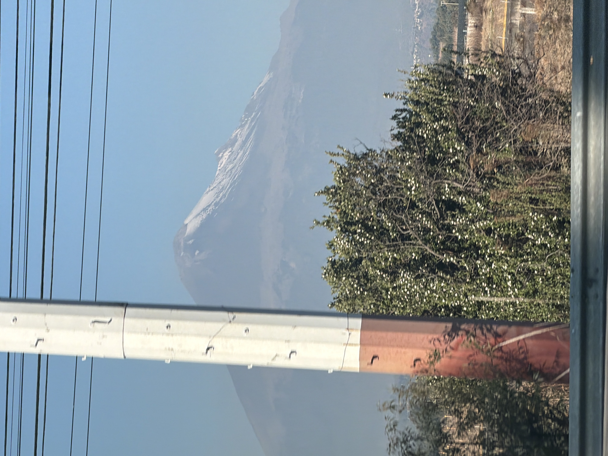The image depicts an outdoor daytime scene in the vicinity of Santa María Zacatepec, Mexico, characterized by a clear sky and distant mountainous terrain.
In the background, a large, snow-capped stratovolcano is visible, partially obscured by atmospheric haze. Its upper slopes are covered in white snow, while the lower slopes appear grey-blue. The sky above and around the mountain is uniformly light blue. Faint, distant residential or urban structures are discernible at the base of the mountain on the far right.
In the midground, a multi-segment utility pole stands vertically, slightly left of the image's center. Its upper section is white/light grey, featuring several small, dark metal attachment hooks. The lower section of the pole transitions to a reddish-brown color. Multiple thin, dark electrical wires run horizontally across the upper portion of the sky, originating from or passing near the pole. To the right of the pole, a dense green bush with numerous small white blossoms occupies a significant portion of the mid-foreground. Sparse, drier brown vegetation is visible to the left of the pole and behind the flowering bush.
In the immediate foreground, a dark grey, metallic guardrail or barrier extends horizontally across the bottom of the frame. No individuals, animals, or specific actions are visible in the scene.