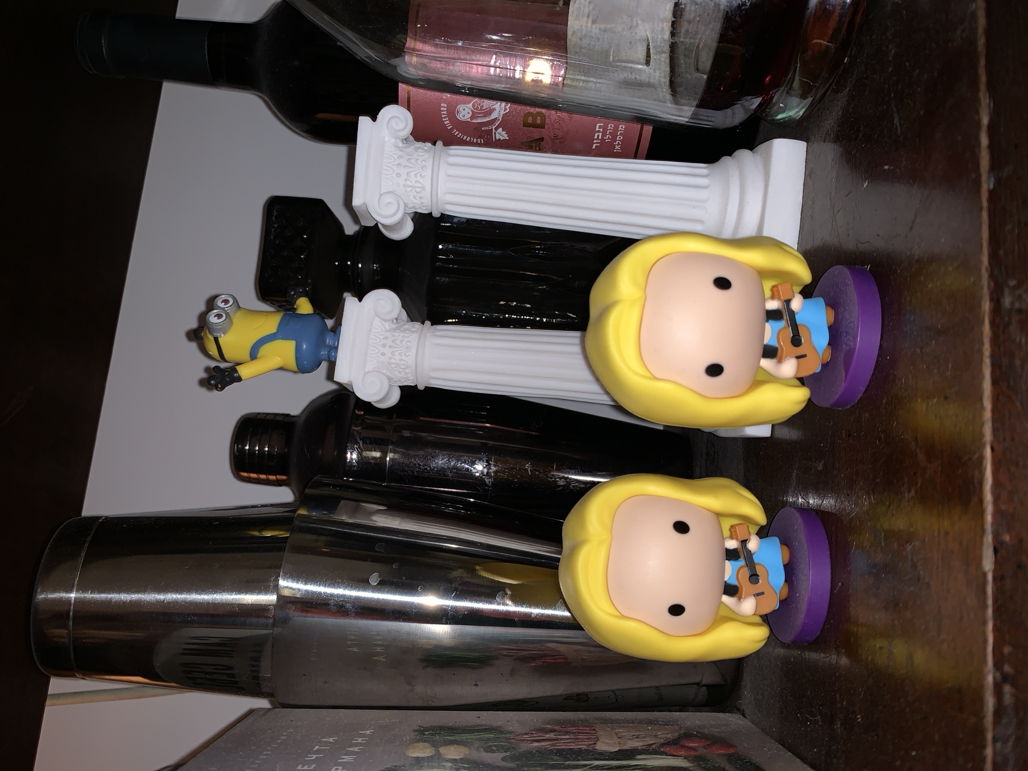 The image displays a cluttered shelf or surface, likely dark wood, presenting a collection of decorative figurines and various bottles. In the foreground, two identical blonde-haired female figurines, resembling Funko Pop characters, are positioned upright on individual purple circular bases. Each figure wears a light blue dress and holds a small brown acoustic guitar.
Behind these, from left to right, a partially visible book or box with a light-colored cover displays text fragments that appear Cyrillic. Adjacent to it, multiple stainless steel cocktail shakers, exhibiting reflections, are stacked. Further right, a yellow Minion figurine with raised arms and goggles stands atop a small white Ionic-style column. Another similar white column is positioned nearby. Behind these columns, a dark green glass wine bottle with a label depicting an owl and the text "ECOLOGICAL VINEYARD" is visible. Below the English text, Hebrew characters reading "יקב אהבה" (Ahava Winery) and "תבור" (Tavor) are present. To the far right, a clear glass decanter or bottle containing a dark amber liquid is partially full. The textual evidence on the wine bottle indicates an Israeli product, placing the scene within a residence or establishment in Tel-Aviv, Israel.