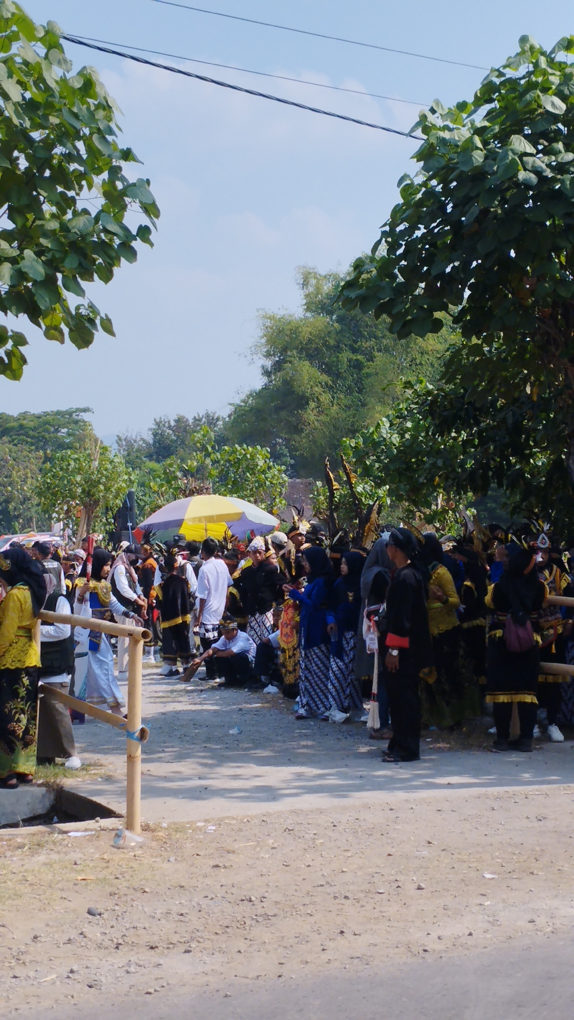 In Bancar, Indonesia, a crowd of people in traditional attire gathers outdoors under a bright, clear sky. Trees with green leaves border the scene, with power lines strung across the top. A yellow and purple umbrella is visible in the background, providing shade for some. Many individuals are dressed in black and gold garments, some with decorative headwear, suggesting a cultural event or festival. The ground is a mix of dirt and gravel, with a small ditch and a wooden fence in the foreground. Some people are standing, while others are seated or kneeling. The atmosphere appears to be lively and communal.