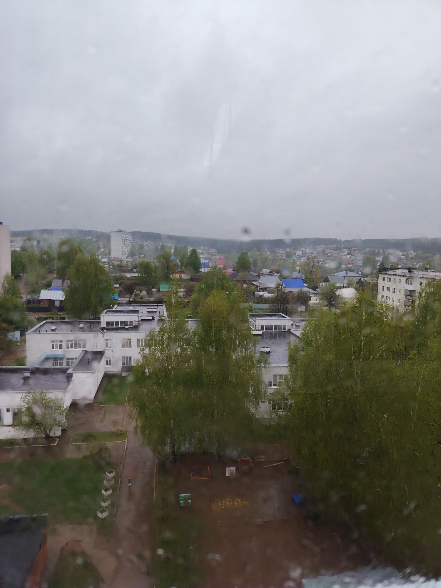 The image shows an elevated view of a residential area in what appears to be Chaikovsky, Russia, taken through a rain-streaked window. Overcast skies and moisture obscure the horizon.
The view encompasses a mix of low-rise buildings, possibly apartments and institutional structures. Several white multi-story buildings with dark roofs are visible amongst a scattering of smaller buildings and houses with blue, red, and grey roofs. Patches of green foliage and trees separate the structures.
A playground area is situated in the foreground, partially obscured by the rain on the window. The presence of playground equipment implies that a kindergarten is nearby. Several large trees are visible in the front yard of the building.