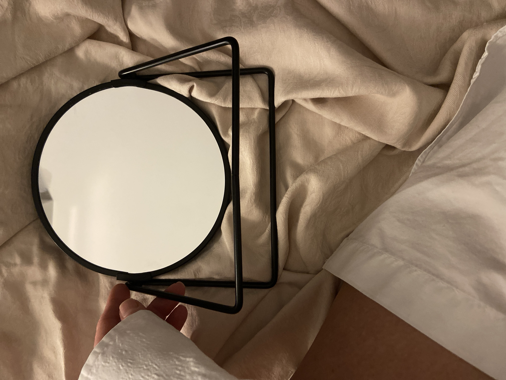 An indoor scene displays a circular tabletop mirror with a black metal frame and stand, resting on a wrinkled light beige fabric surface with a subtle woven pattern. A segment of an arm, covered by a white fabric sleeve, extends from the left foreground, with an exposed area of bare skin visible below the sleeve. A hand from this arm is in contact with the upper left section of the mirror's black frame. The mirror's reflective surface shows a bright, indistinct white light source. The precise location within the city of Budva, Montenegro, is not discernible from the visual information provided.