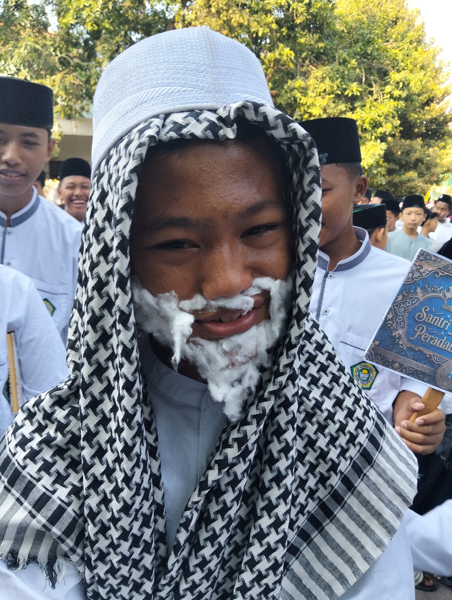A close-up, eye-level shot shows a young male in Kranji, Indonesia, wearing a white kufi and a black and white keffiyeh draped around his head. A white substance, possibly shaving cream or cotton, is applied to his upper lip and chin, resembling a beard. He is smiling and looking towards the camera. In the background, other individuals, mostly young males in white attire and kufis, are visible. One person to the right is holding a book or sign with blue and gold lettering. The scene appears to be outdoors, with trees and foliage in the background, suggesting a bright, sunny day. The image is sharp and well-lit, focusing on the central subject and his playful application of the white substance.