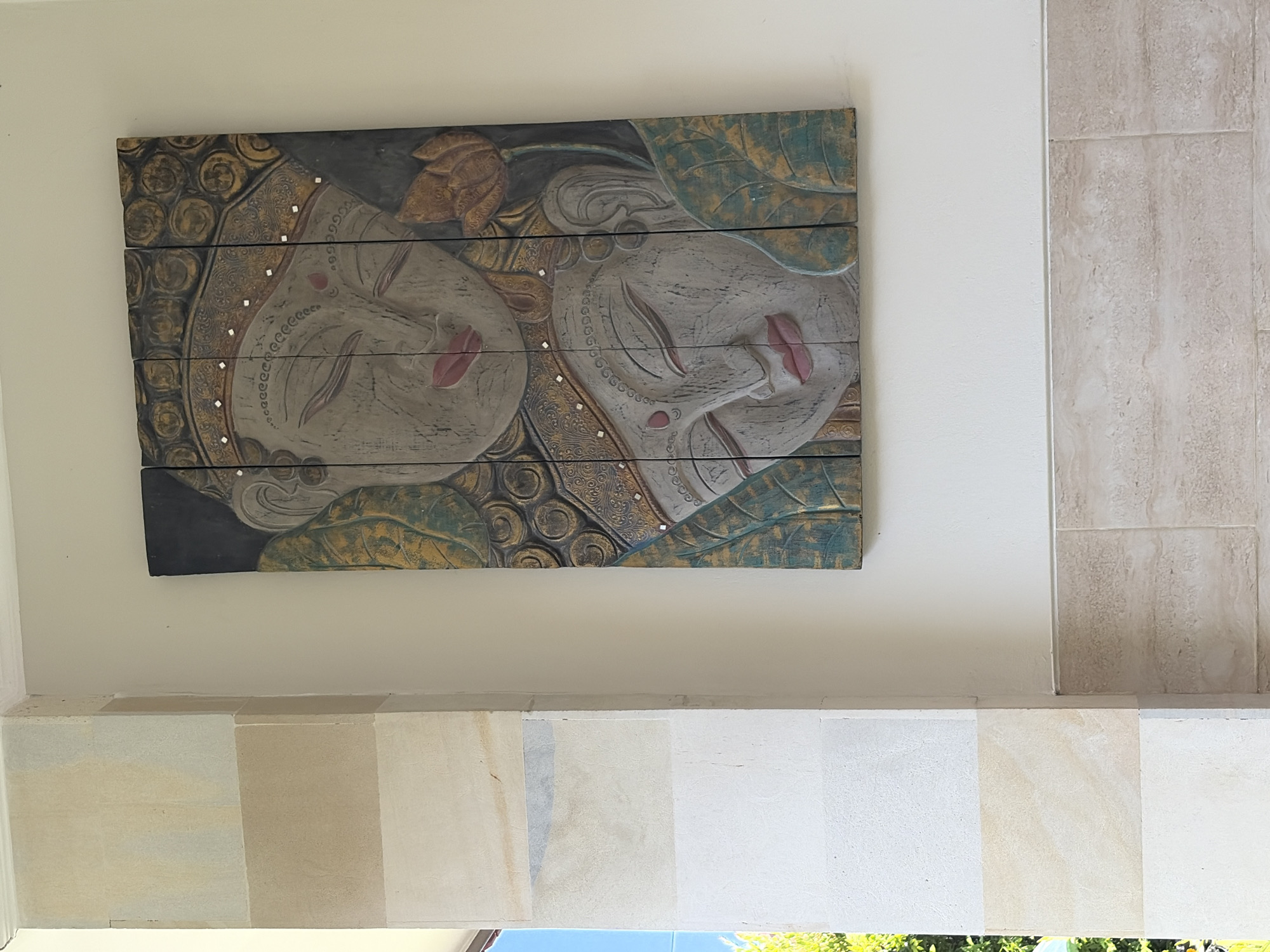 The image shows a carved wooden panel depicting two serene faces with closed eyes, possibly of Buddha or a similar spiritual figure. The panel is mounted on a light-colored wall, which is slightly textured. To the left of the panel is a structural pillar clad in large, rectangular stone tiles of varying beige and off-white tones. Below the panel and extending across the bottom of the image is a tiled section of the wall, featuring smaller, horizontally laid beige tiles with a marbled texture. Natural light suggests an outdoor or semi-outdoor setting, possibly a building facade or veranda. The location is Kuta Selatan, Indonesia, a region known for its rich cultural heritage and artistic traditions, which is reflected in the style of the artwork.