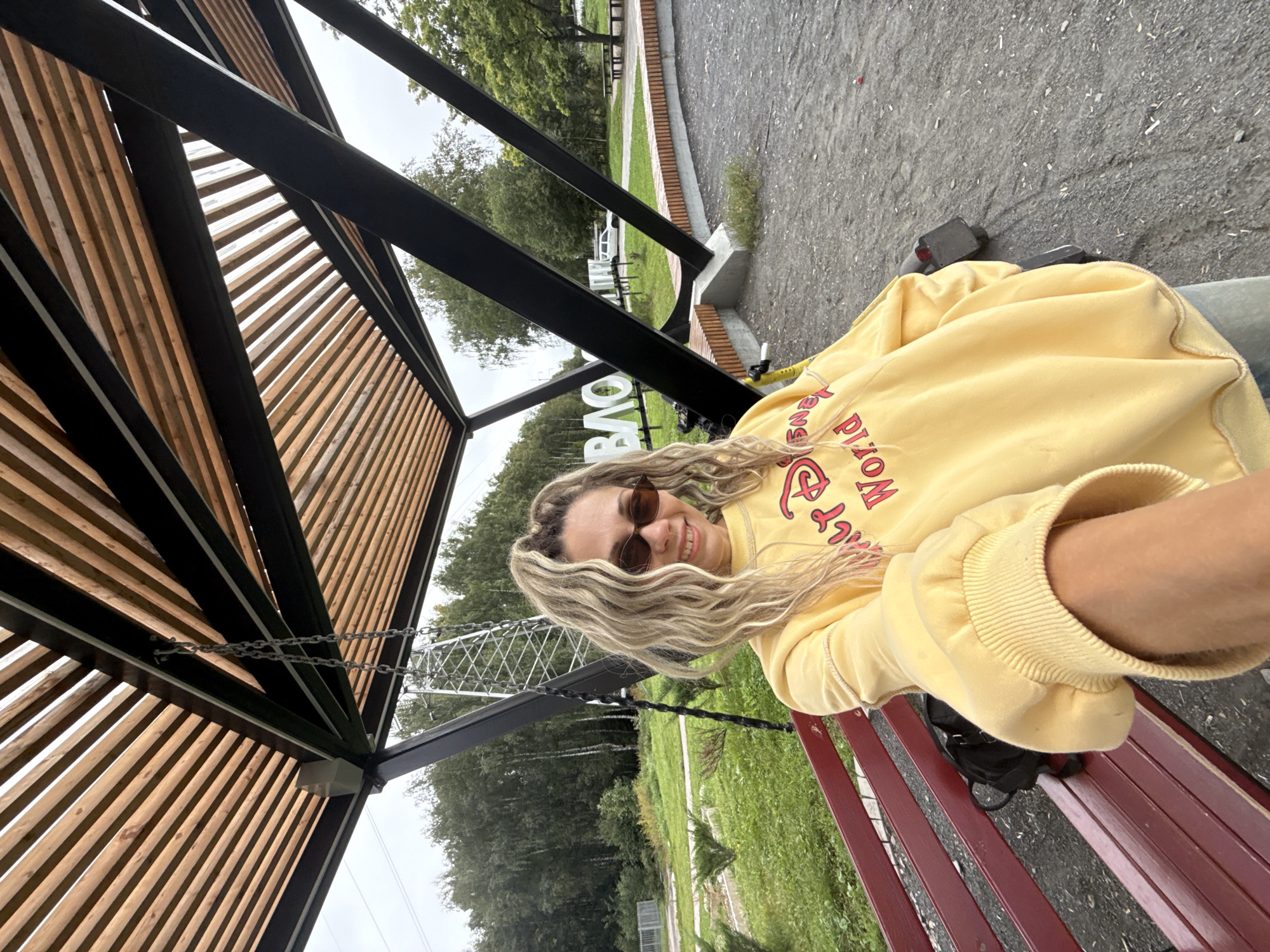 In Rusanovo, Russia, a woman with blonde, wavy hair and sunglasses is taking a selfie while sitting on a red bench. She is wearing a yellow sweatshirt with "Disney World" written on it in red lettering. She is positioned under a modern wooden and metal structure, possibly a pavilion or shelter, with a swing set visible behind her. The background consists of a park-like setting with trees, grass, and a large sign with white letters spelling out "ВАО". A white car is parked in the distance, and the sky is overcast. The ground around the bench is covered with gravel and dirt.