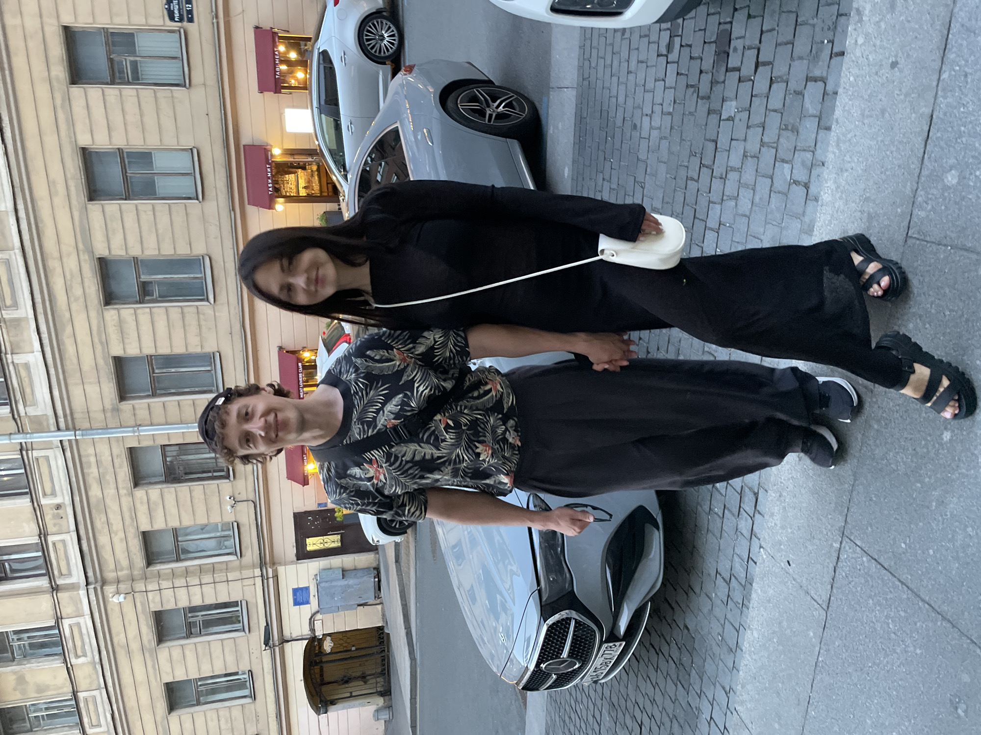 The image captures a full-length portrait of a man and a woman holding hands on a street in Saint Petersburg, Russia. The location is identifiable as Rubinshteina Street, specifically near number 12, as indicated by a visible street sign and the "ТАБИ.МИТ" (Tabi Meat) restaurant signage on the ground floor building.
The man, positioned on the left, has light skin, short curly brown hair, and wears a dark baseball cap backward. He is smiling and looking directly at the camera. He is wearing a dark short-sleeved shirt with a tropical print featuring green leaves and red-orange flowers, dark loose-fitting trousers, and dark sneakers with white soles. A black crossbody bag with a wide strap is slung over his shoulder. He holds a pair of glasses in his right hand.
The woman, positioned on the right, has light skin, long straight dark hair parted in the middle, and is also smiling slightly while looking directly at the camera. She is wearing a long-sleeved, boat-neck, black maxi dress and black chunky-soled sandals. A small white crossbody bag with a thin strap hangs over her left shoulder. Her left hand is holding the man's right hand.
They are standing on a light grey paved sidewalk. Behind them, the street is paved with grey cobblestones/pavers. A light grey Mercedes-Benz sedan with a partially visible Russian license plate "H158EX 778" is parked directly behind the man. Further back, a silver Kia Optima sedan is also parked.
The background features a multi-story building with a light beige facade, numerous windows, and decorative architectural elements. The ground floor houses commercial establishments, including "ТАБИ.МИТ" (Tabi Meat) and "MEAT LOVERS CLUB," visible under red awnings. A blue square sign and a grey electrical box are visible on the building wall to the left of the restaurant entrance. A grey street lamp pole runs vertically in the mid-ground.