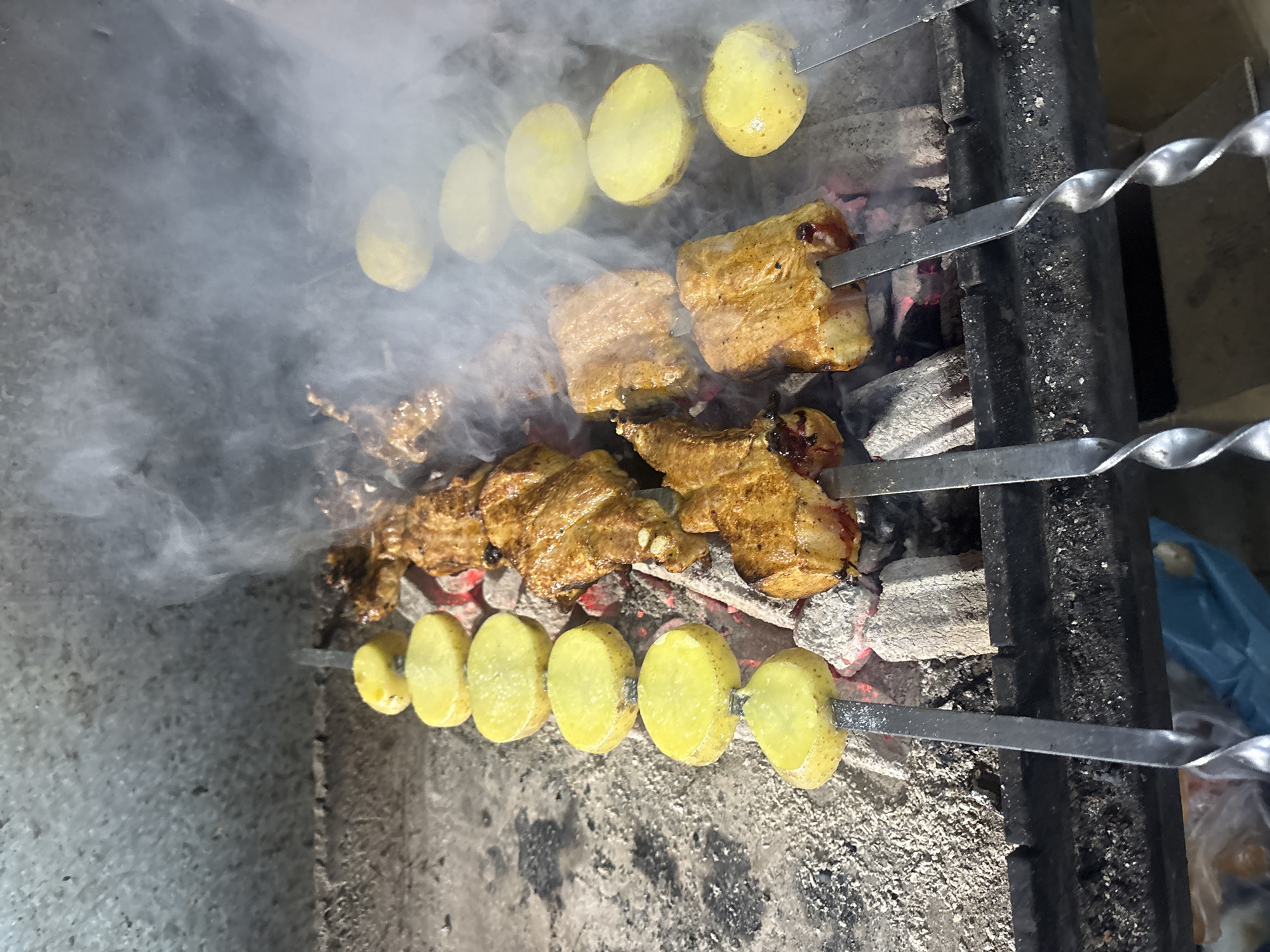 The image depicts skewers of meat and potatoes being grilled over hot coals. Visible is dense smoke rising from the cooking food. The skewers are supported by a metal frame over a bed of gray coals. The setting is outdoors, possibly a backyard or park, but the specific location is not discernible from the image. There are no people visible. There is a blue bag that is visible in the background.