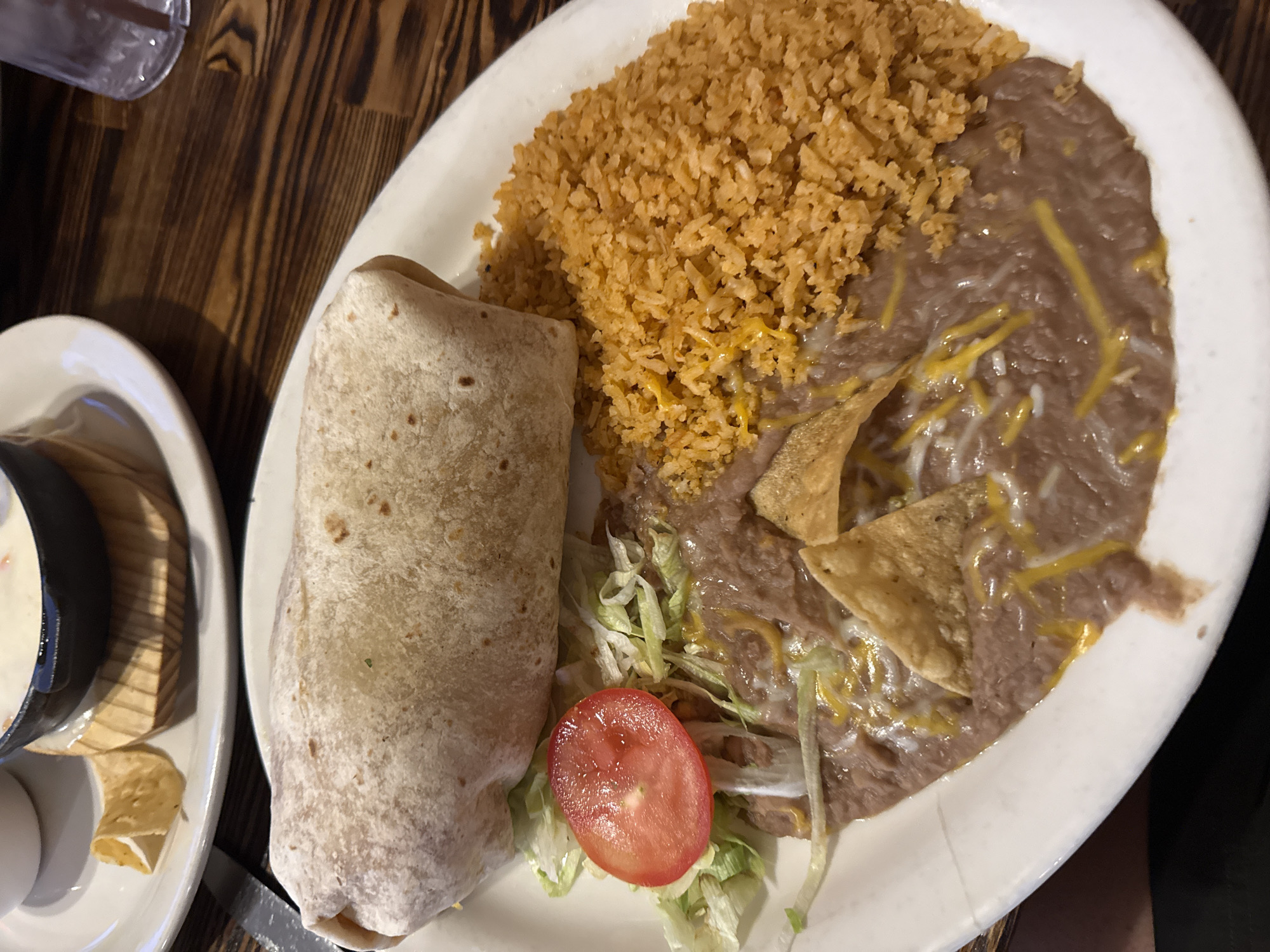 A high-angle shot shows a white plate of Mexican food on a dark wooden table, likely inside a restaurant. The plate contains a large burrito, a mound of orange rice, and a portion of refried beans topped with shredded cheese and tortilla chips. A small pile of shredded lettuce and a slice of tomato garnish the plate. To the left of the plate is a white saucer holding a small, dark bowl filled with a white, creamy substance, possibly cheese dip or sour cream, accompanied by a few tortilla chips. A drinking glass is partially visible in the upper-left corner of the frame. There is no visible indication of the location in Palm Springs, United States of America.