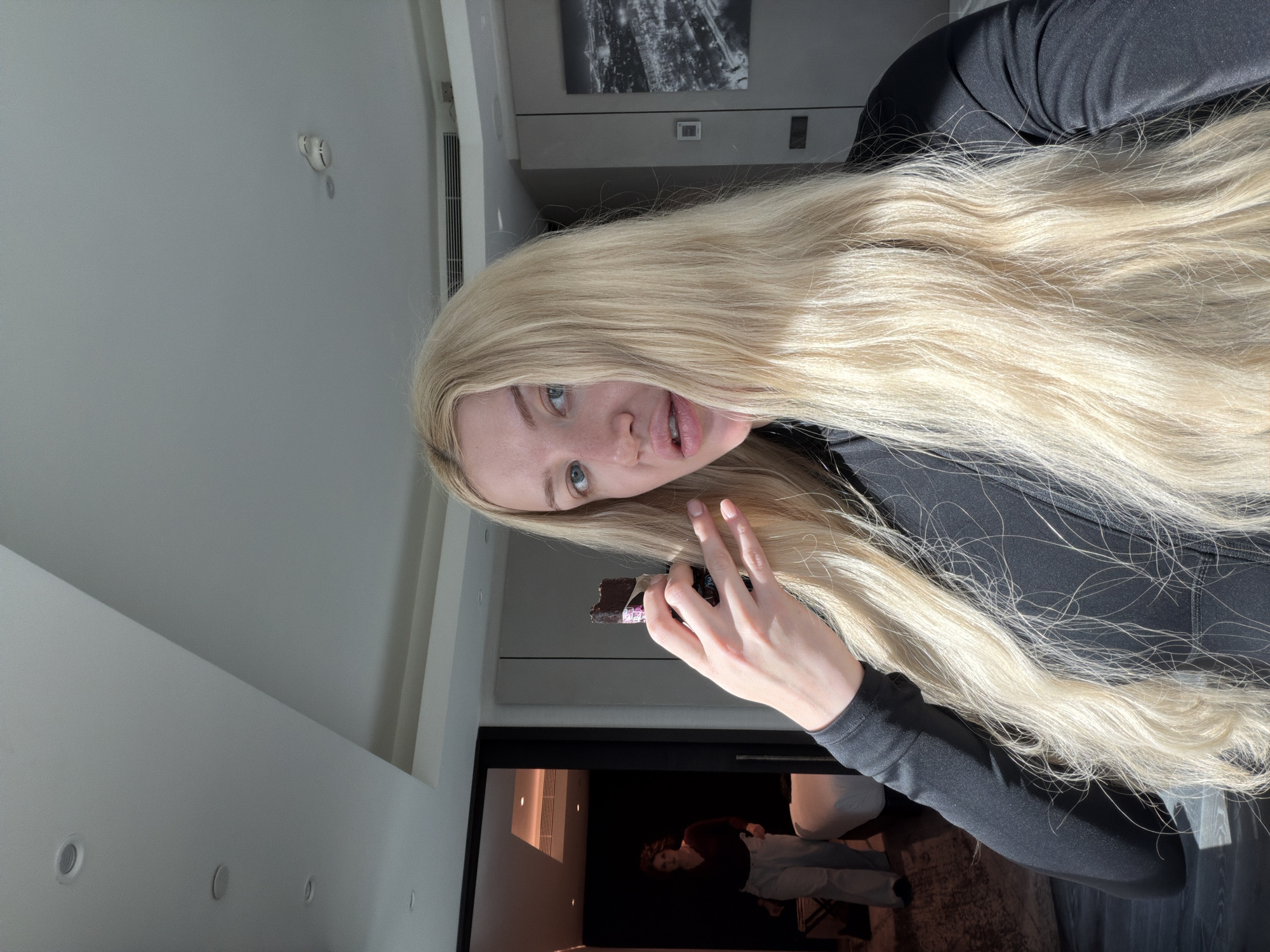 A woman with long blonde hair is holding a partially unwrapped chocolate bar in her right hand. She is wearing a black, long-sleeved shirt. In the background, through a doorway, a person is visible wearing a maroon top and light-colored pants. The room appears to be interior space with light-colored walls and recessed lighting. A piece of framed art is seen on the wall near the ceiling. The location and the city of Vracar Urban Municipality, Serbia, is unidentifiable.